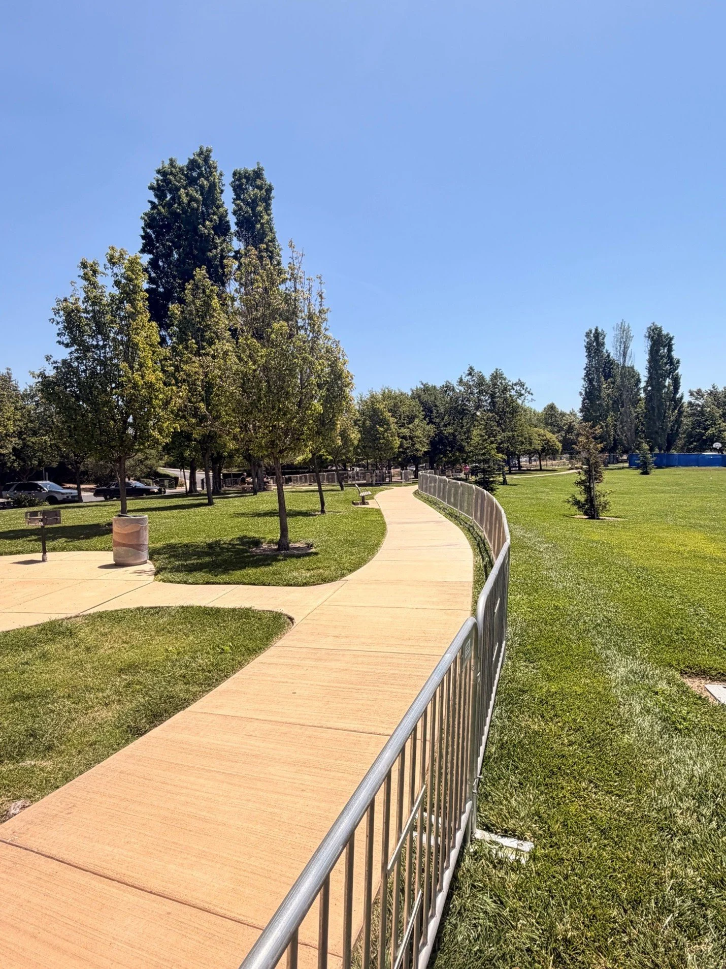 A park with a curved concrete walking path, green grass, several trees, benches, and a metal fence, under a clear blue sky.