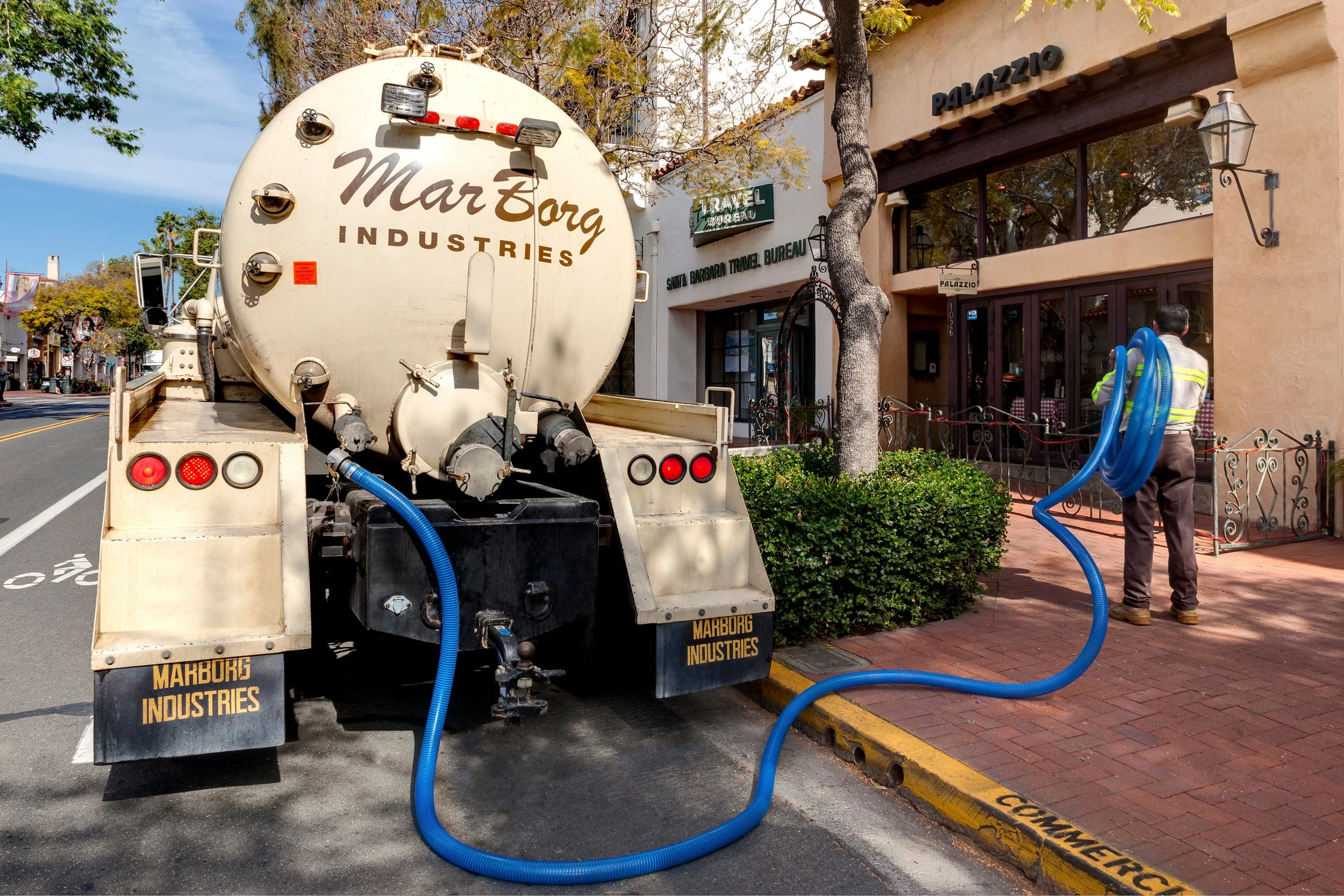 A street scene with a large white tanker truck labeled 'Marborg Industries' parked next to a sidewalk. The truck has blue hoses connected to the back, and a worker is visible holding more hoses. The background features storefronts, trees, and a clear sky.