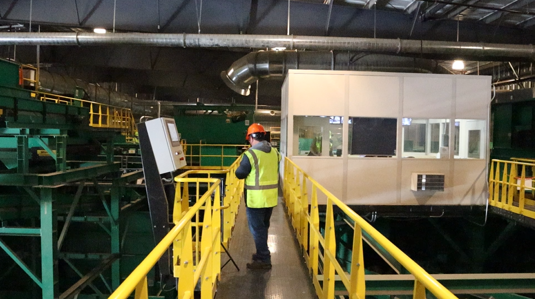 Worker in safety gear on a yellow metal walkway inside an industrial facility with large machinery and ventilation ducts.