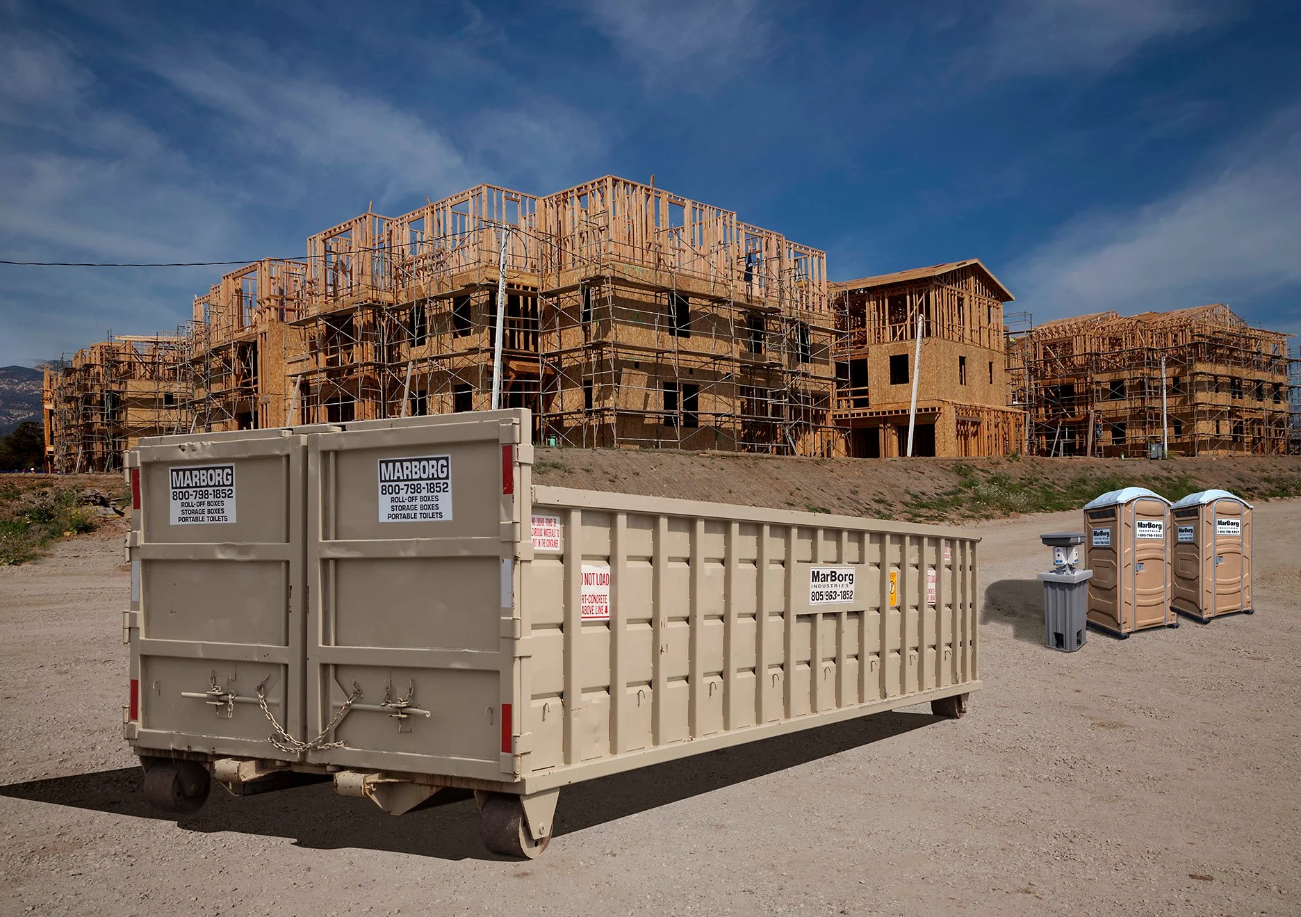 Construction site with a multi-story wooden building framework surrounded by scaffolding. In the foreground, there are large beige containers labeled 'Marborg' and portable toilets. The sky is clear with some clouds, and the area appears to be in a developing residential or commercial area.