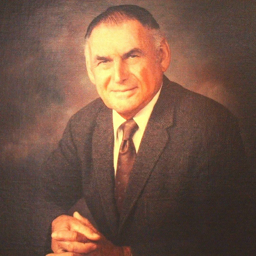 Portrait of Mario Borgatello in a suit with a tie, sitting with arms crossed, smiling slightly, against a dark background.