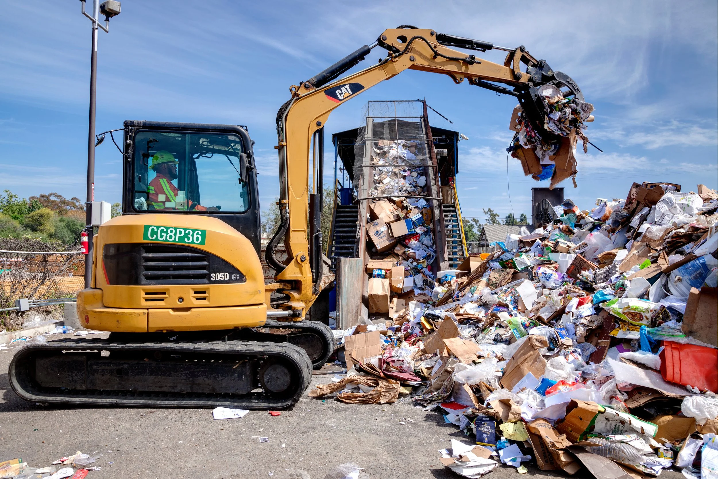 A yellow CAT excavator is moving a large pile of mixed garbage, including cardboard, paper, and plastic, in an outdoor landfill site under a partly cloudy sky.