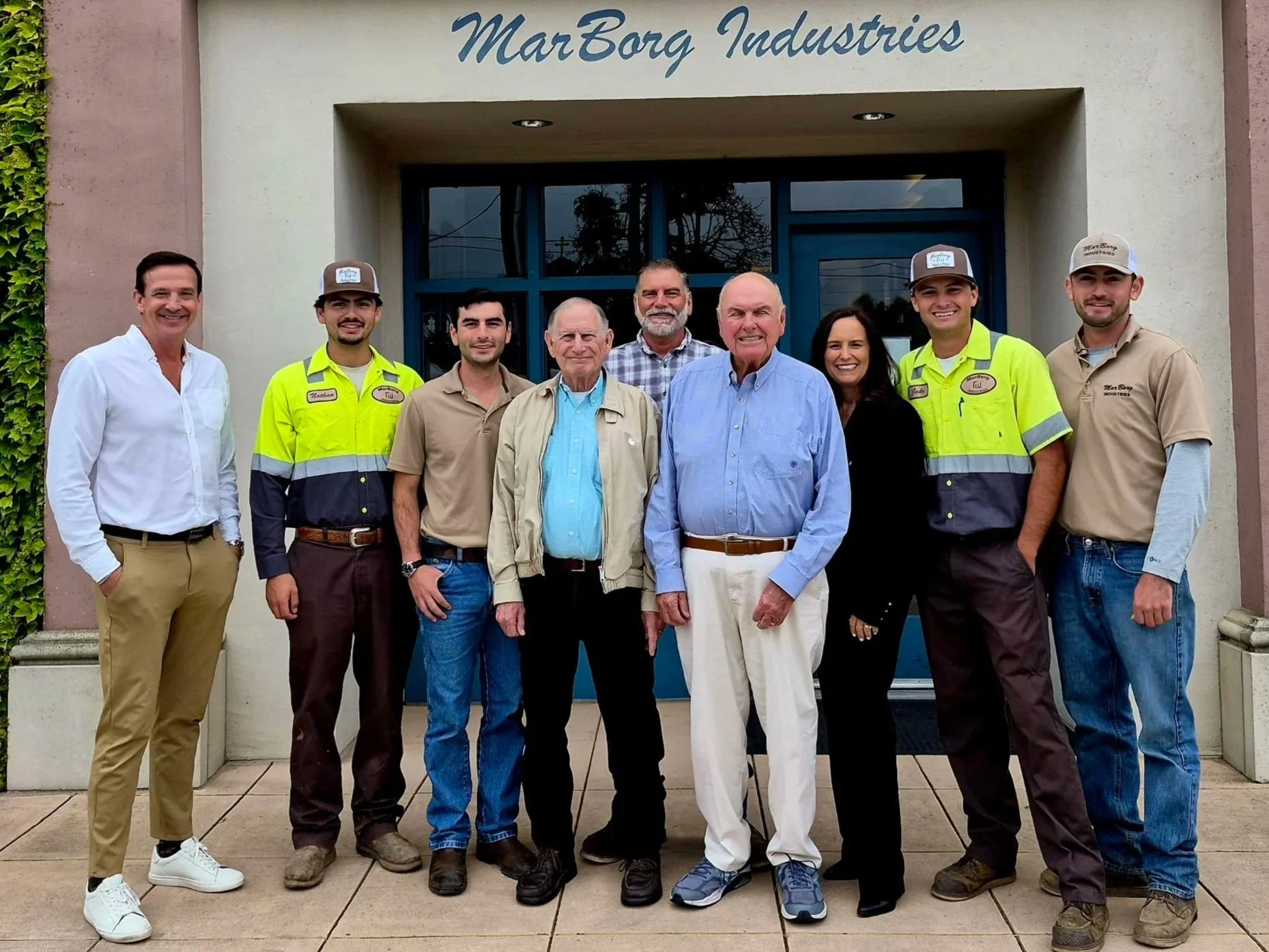 A group of nine people standing outside the entrance to MarBorg Industries, smiling for the photo. The group includes men and women of various ages, some in casual clothes and a few in work uniforms. The background shows a building with the company name above the entrance.