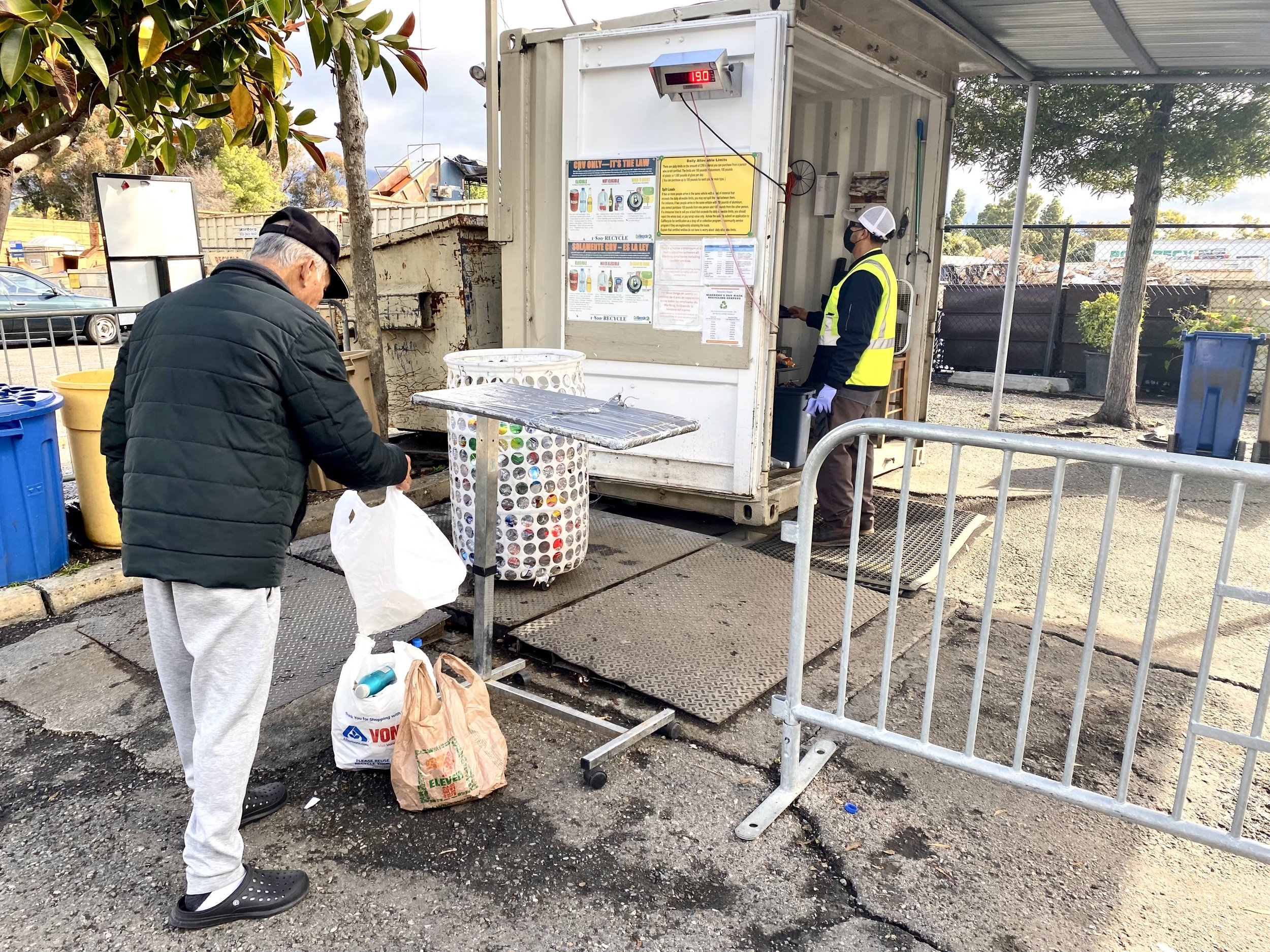 A man with gray hair wearing a black cap, black jacket, and gray sweatpants standing at a compost bin outside of a recycling center, with his back to the camera. There is a young worker in a high-visibility vest and face mask handling paperwork at the recycling station. Several bags of recyclables, including one with a 7-Eleven logo, are on the ground. The recycling station has various informational posters and a digital display showing the temperature of 19.0 degrees Celsius.