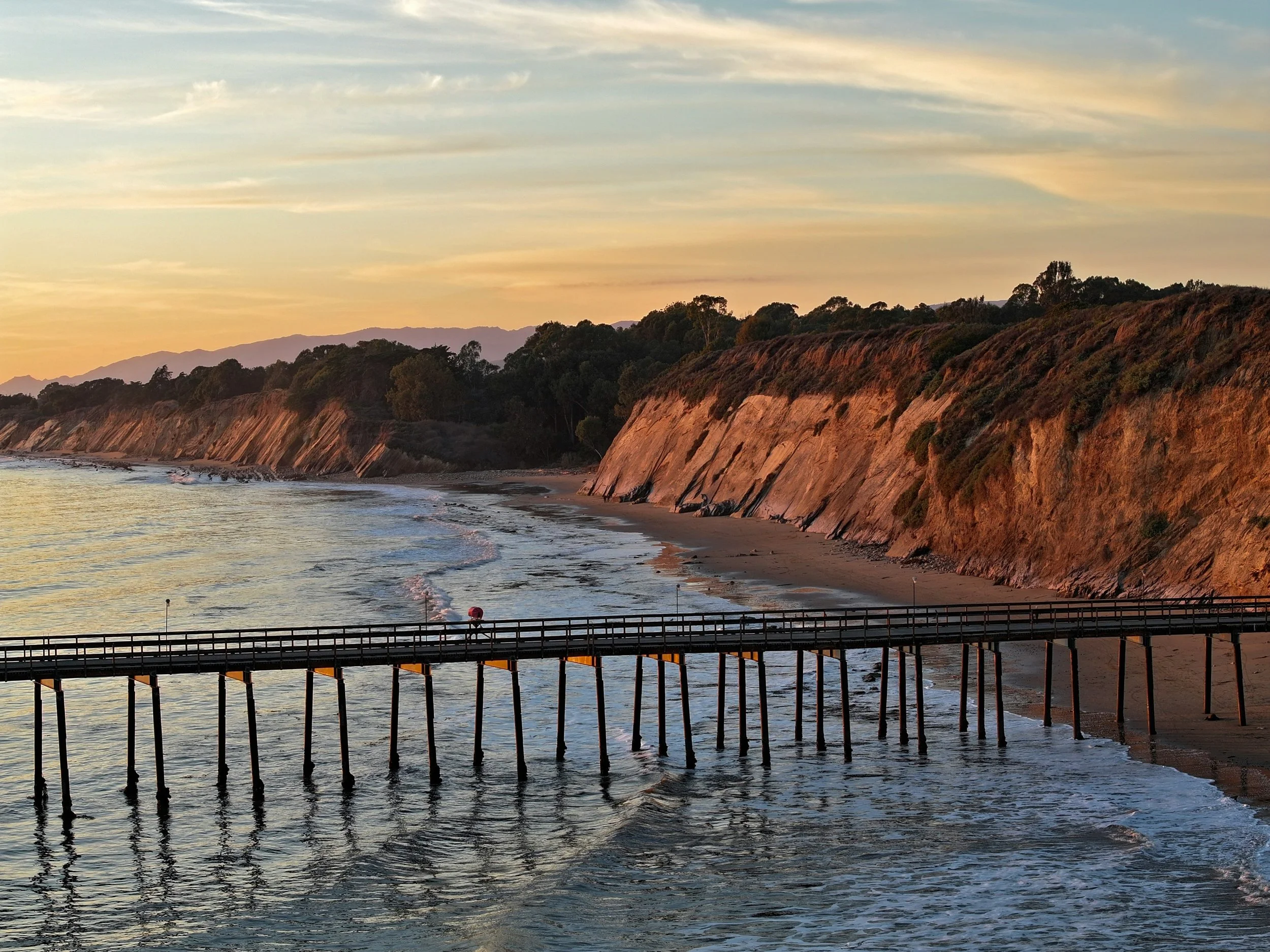 Sunset over the Goleta coastline with a wooden pier extending into the water, cliffs with trees beyond, and mountains in the background.