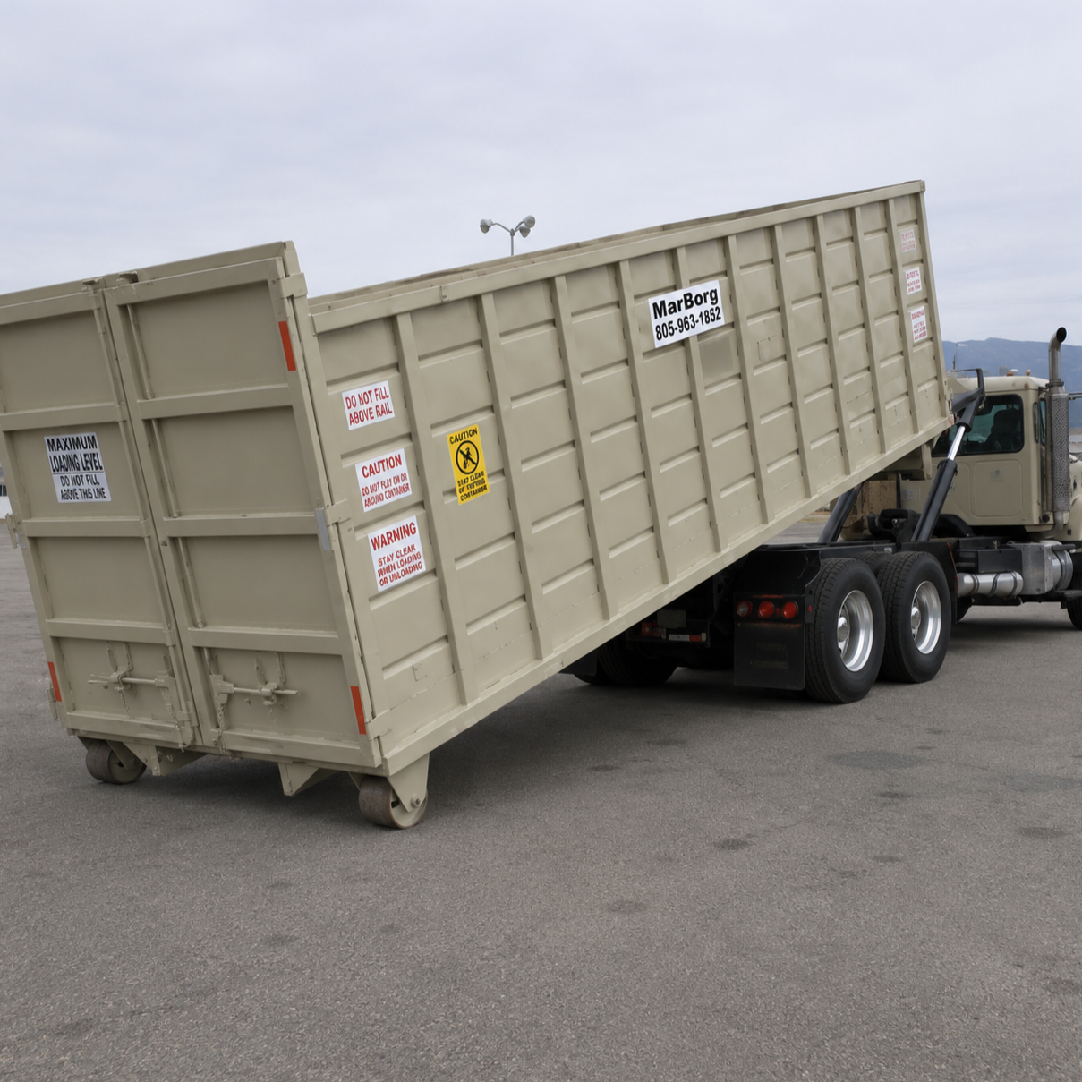 A large beige roll-off container, being set on a paved area outdoors with mountains and trees in the background.