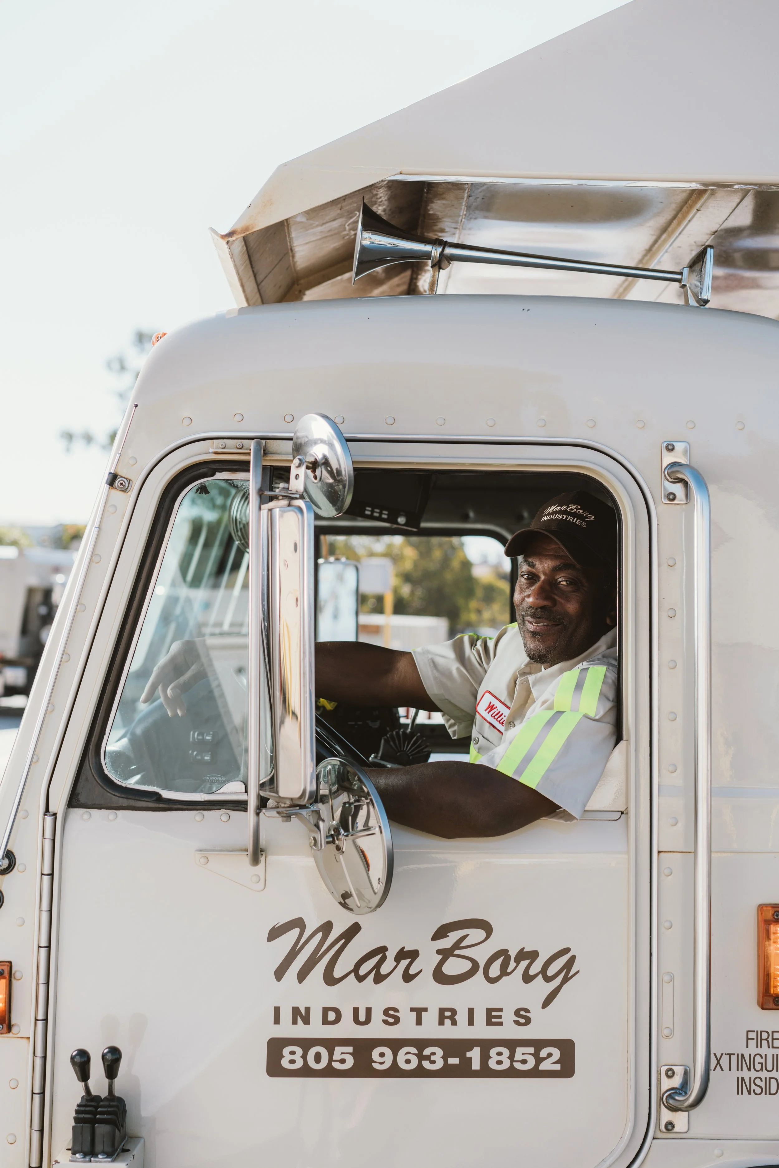 A man in a white uniform with yellow reflective stripes, wearing a cap with the words 'Willi Co' and 'Industries,' is sitting in the driver's seat of a white food truck called 'Mar Borg Industries.' The truck has a large side mirror and various controls visible on the door and dashboard. The background shows trees and sky.