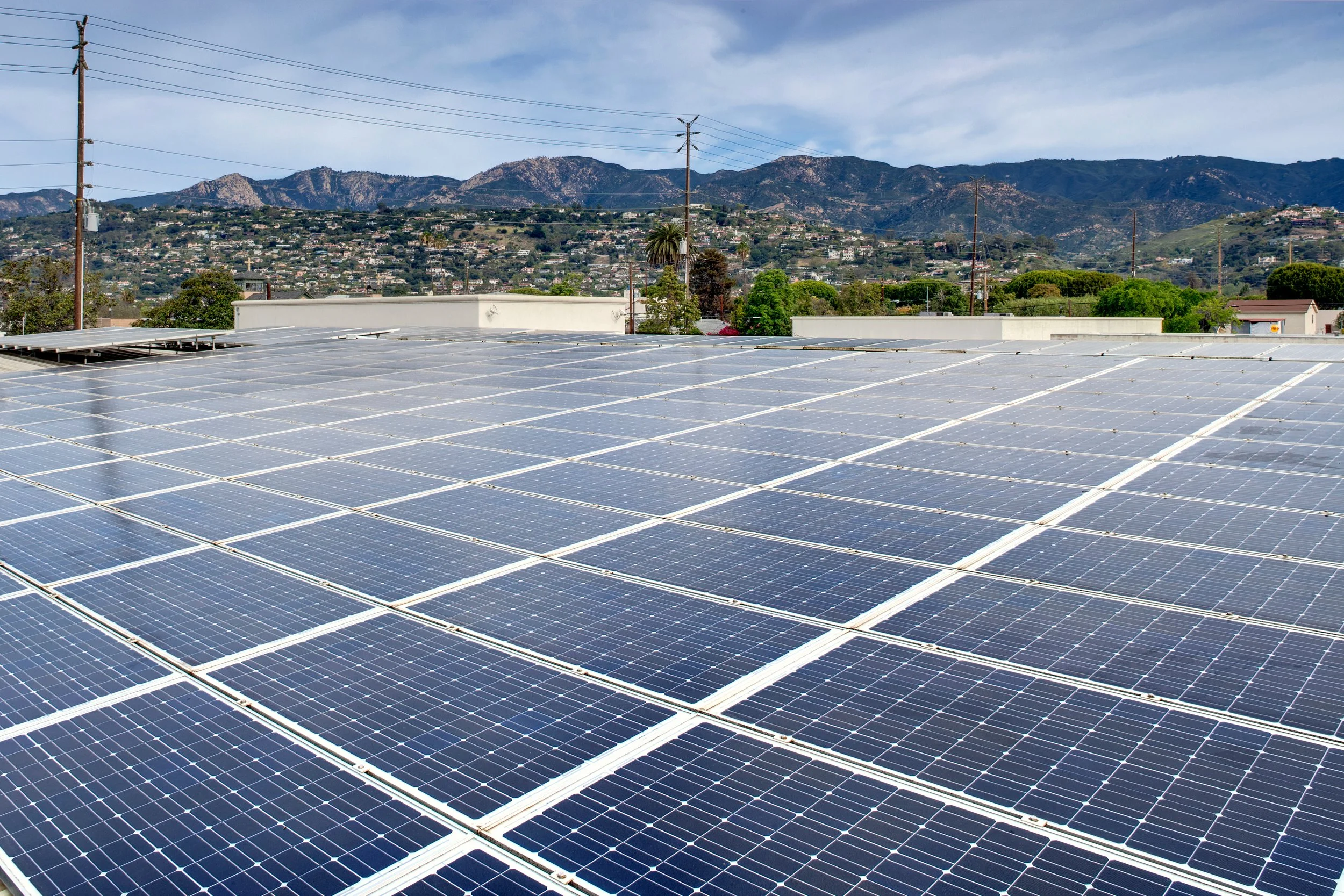 A large array of solar panels installed on a rooftop with a scenic view of hills and houses in the background on a partly cloudy day.
