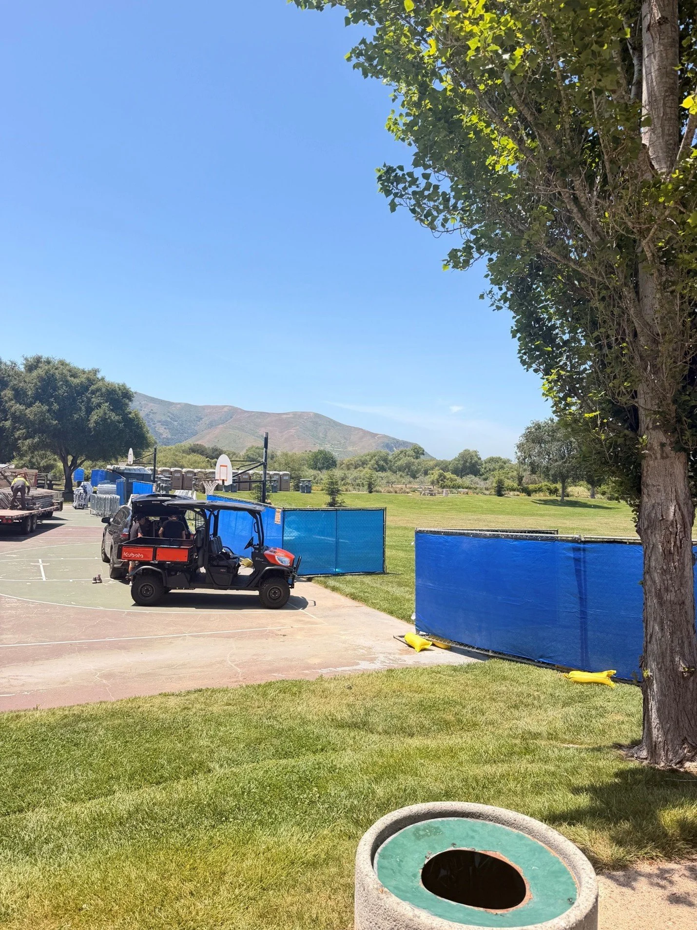 A park with a grassy area, trees, and mountains in the background. There is a basketball court with benches, a golf cart, and a few people in the distance. Blue barriers divide part of the area, and a trash bin in the foreground.