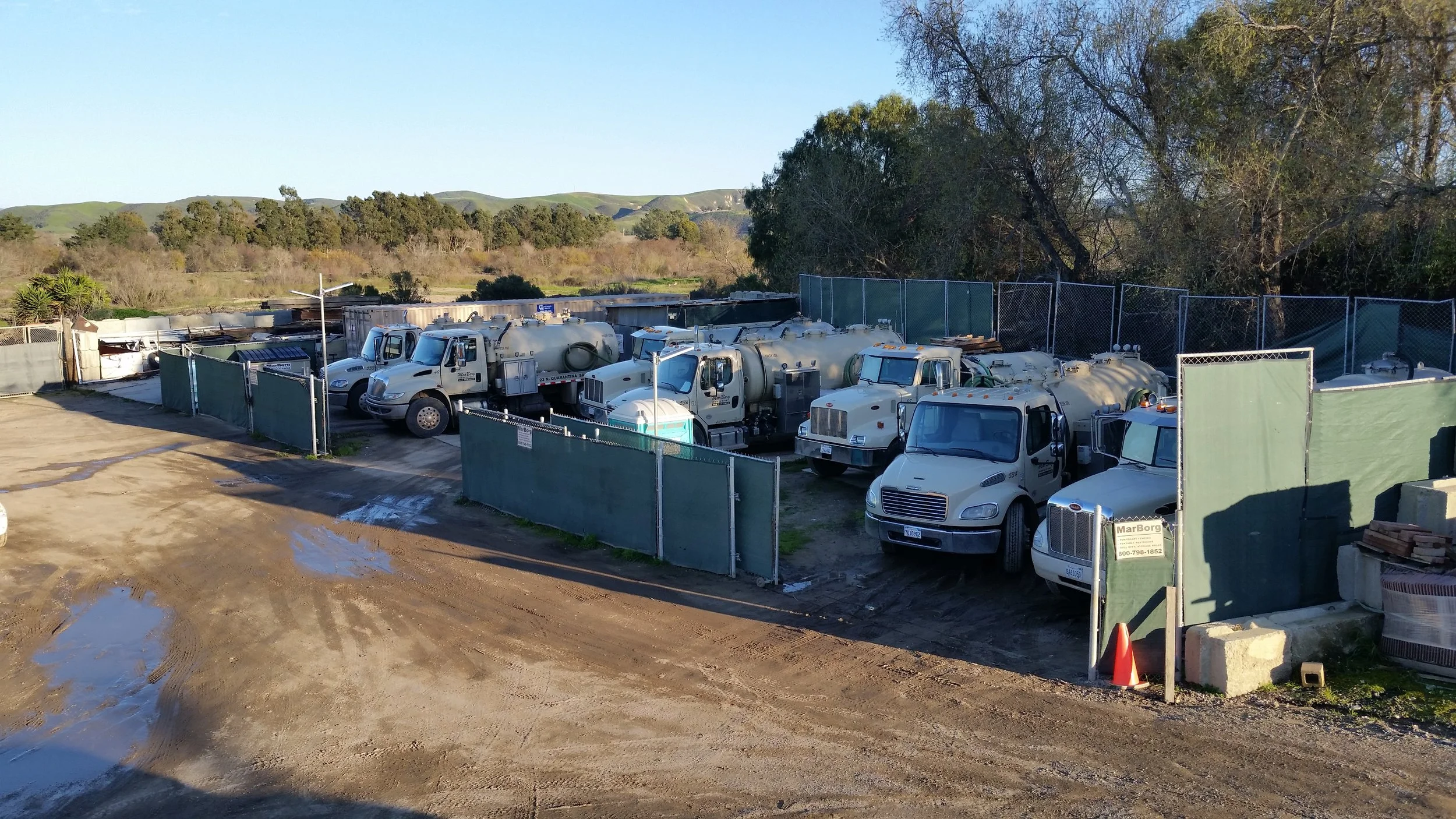 A fenced lot containing several water trucks and vans, with a dirt ground and puddles, surrounding trees, and green hills in the background.