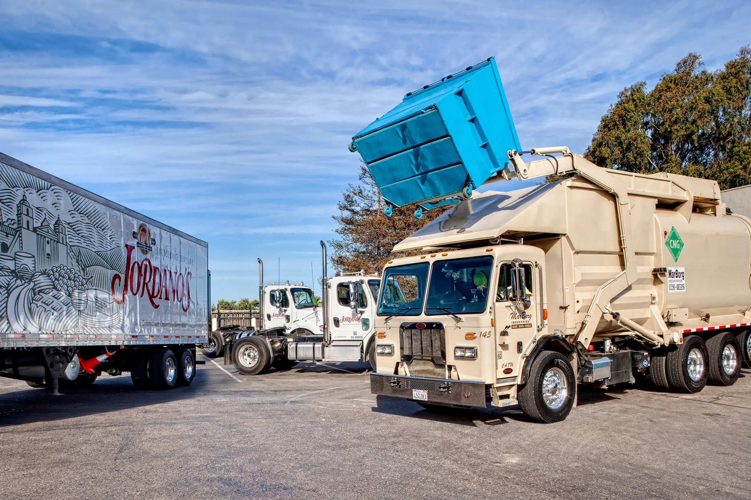 A line of trucks parked in a lot, with one truck's hydraulic arm lifting a blue dumpster into the air. The sky is partly cloudy, and there are trees and a billboard in the background.