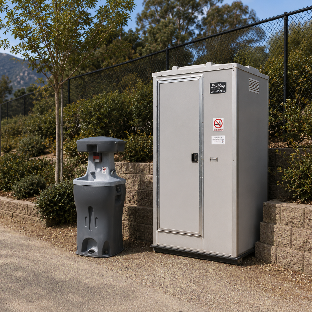 Utility box and trash bin on sidewalk with a chain-link fence and trees in the background.