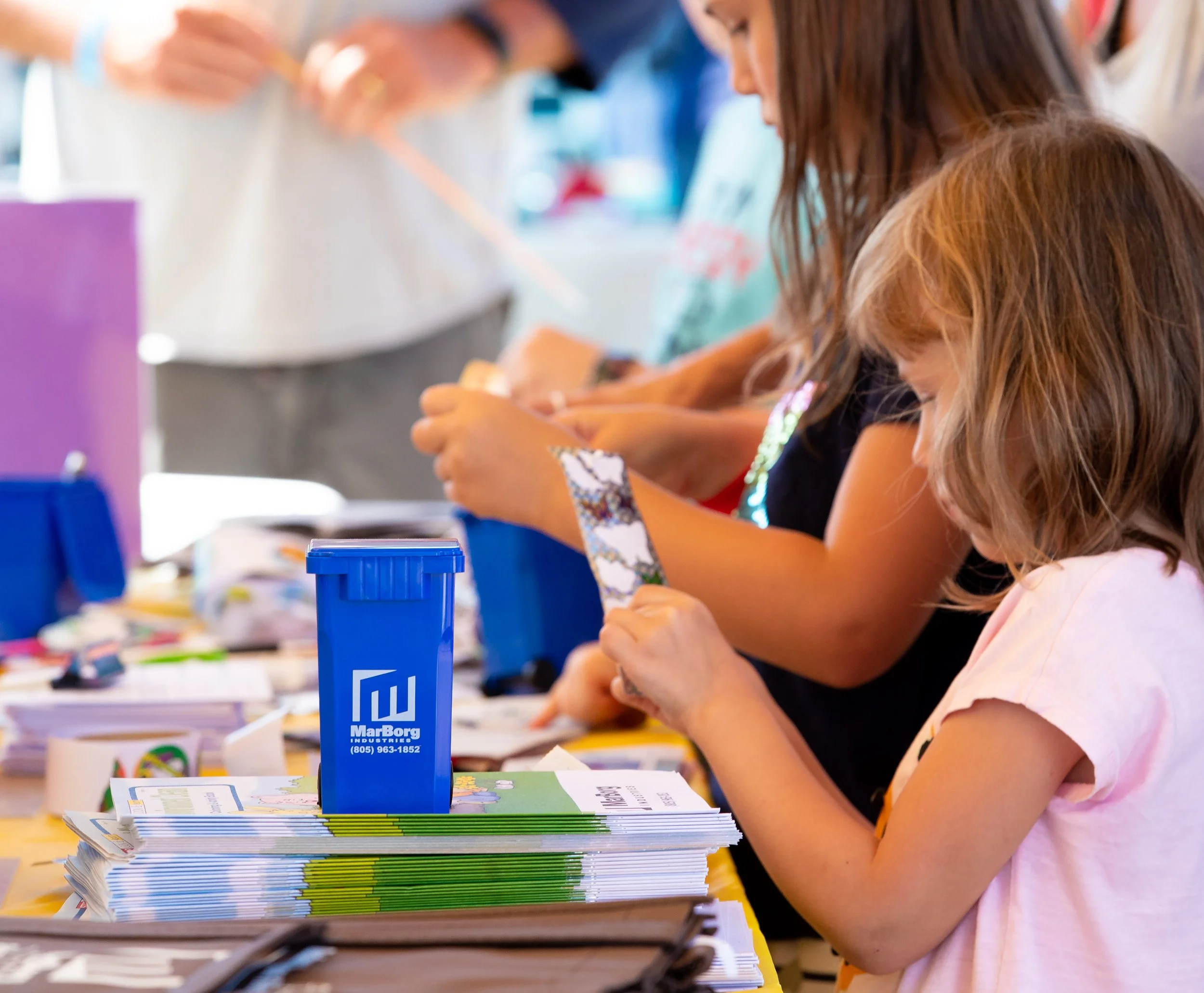 Children sitting at a table, engaged in an activity, with colorful books, pamphlets, and a blue MarBorg container on the table.