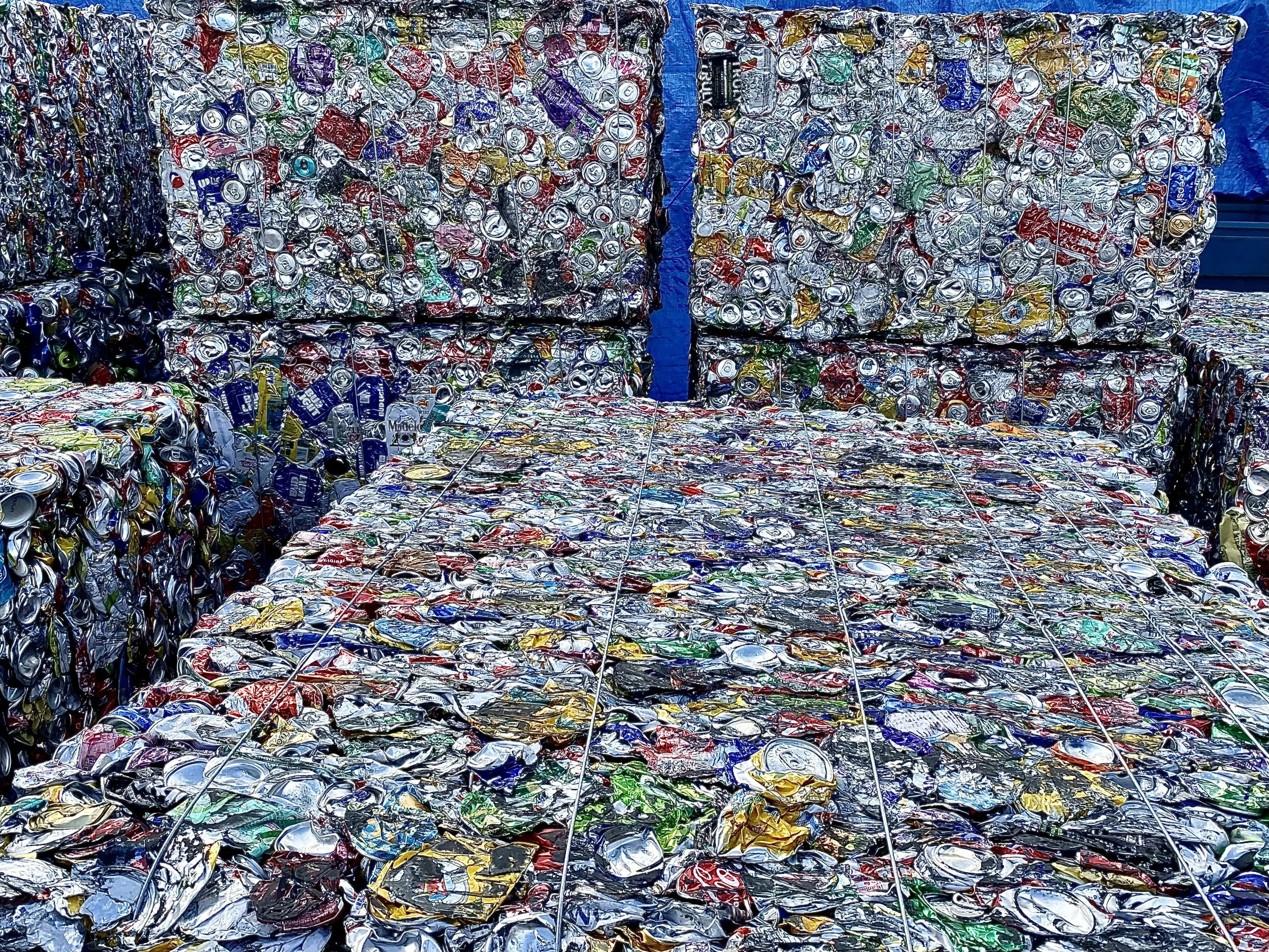 Large piles of crushed aluminum cans are stacked in an outdoor recycling area, with some blue tarps and metal fencing visible in the background.
