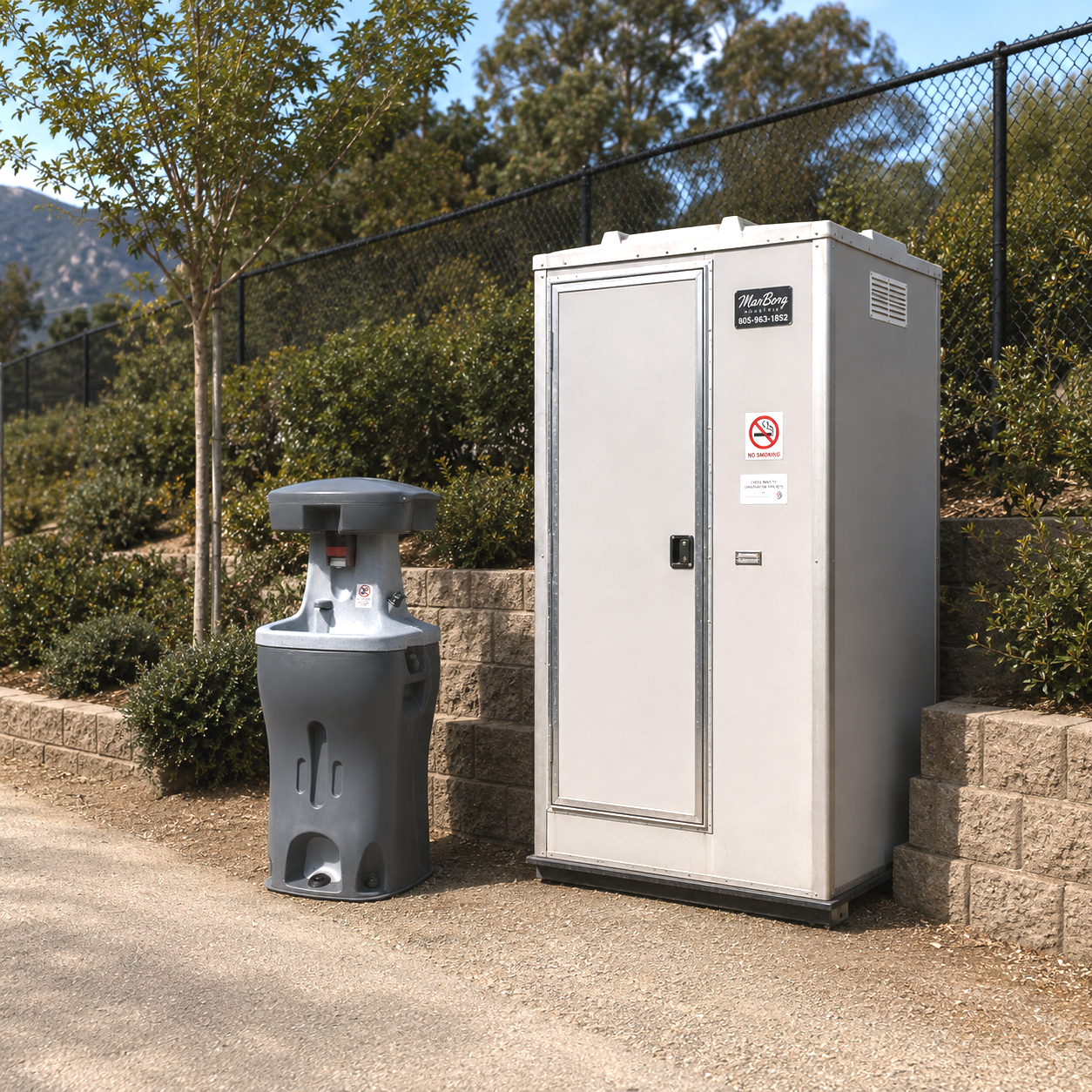 Public outdoor trash and recycling containers next to a landscaped area with bushes and trees, enclosed by a black chain-link fence, in a sunny setting.