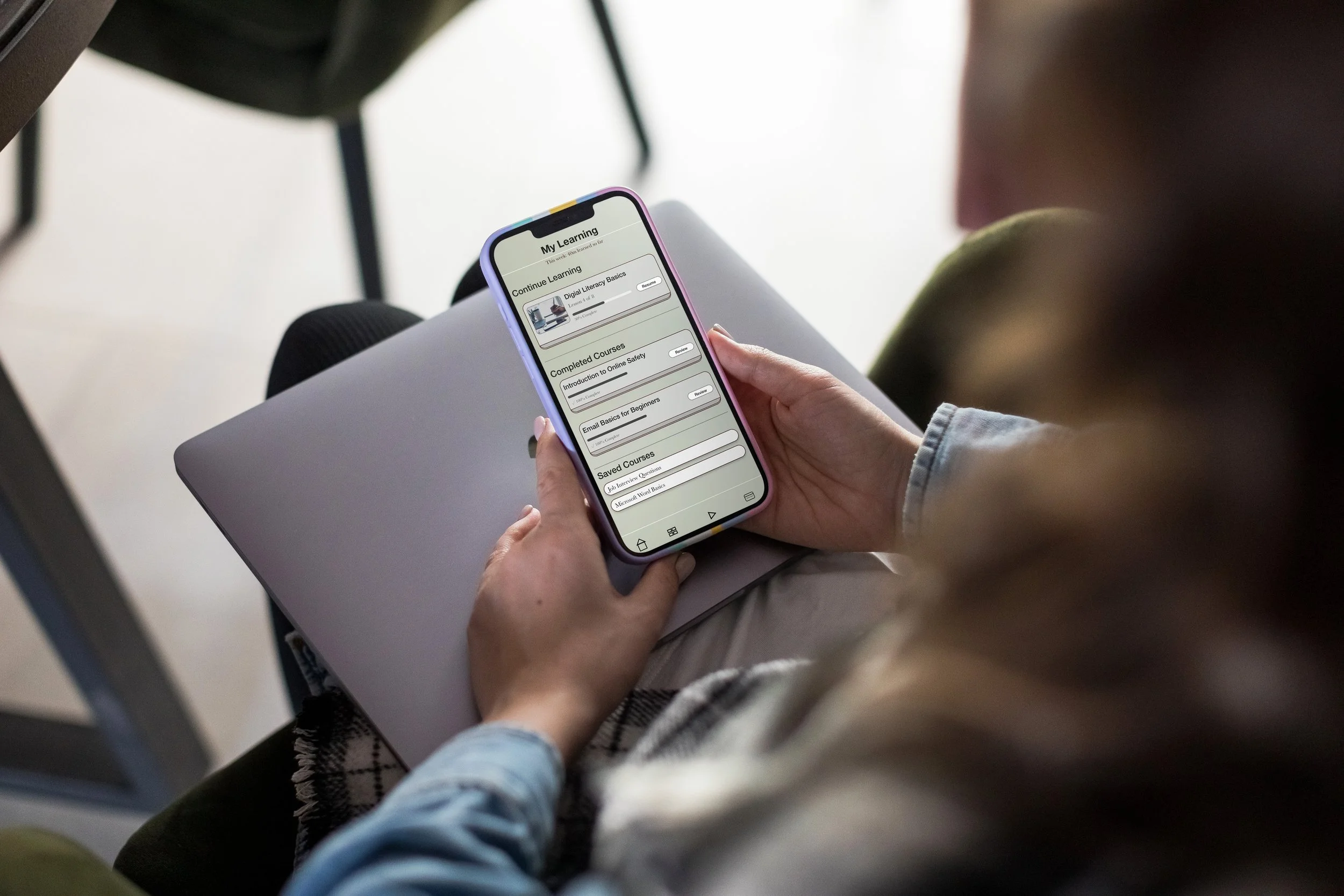 Person holding a smartphone with an educational app open, sitting with legs crossed on a gray surface, near a chair.