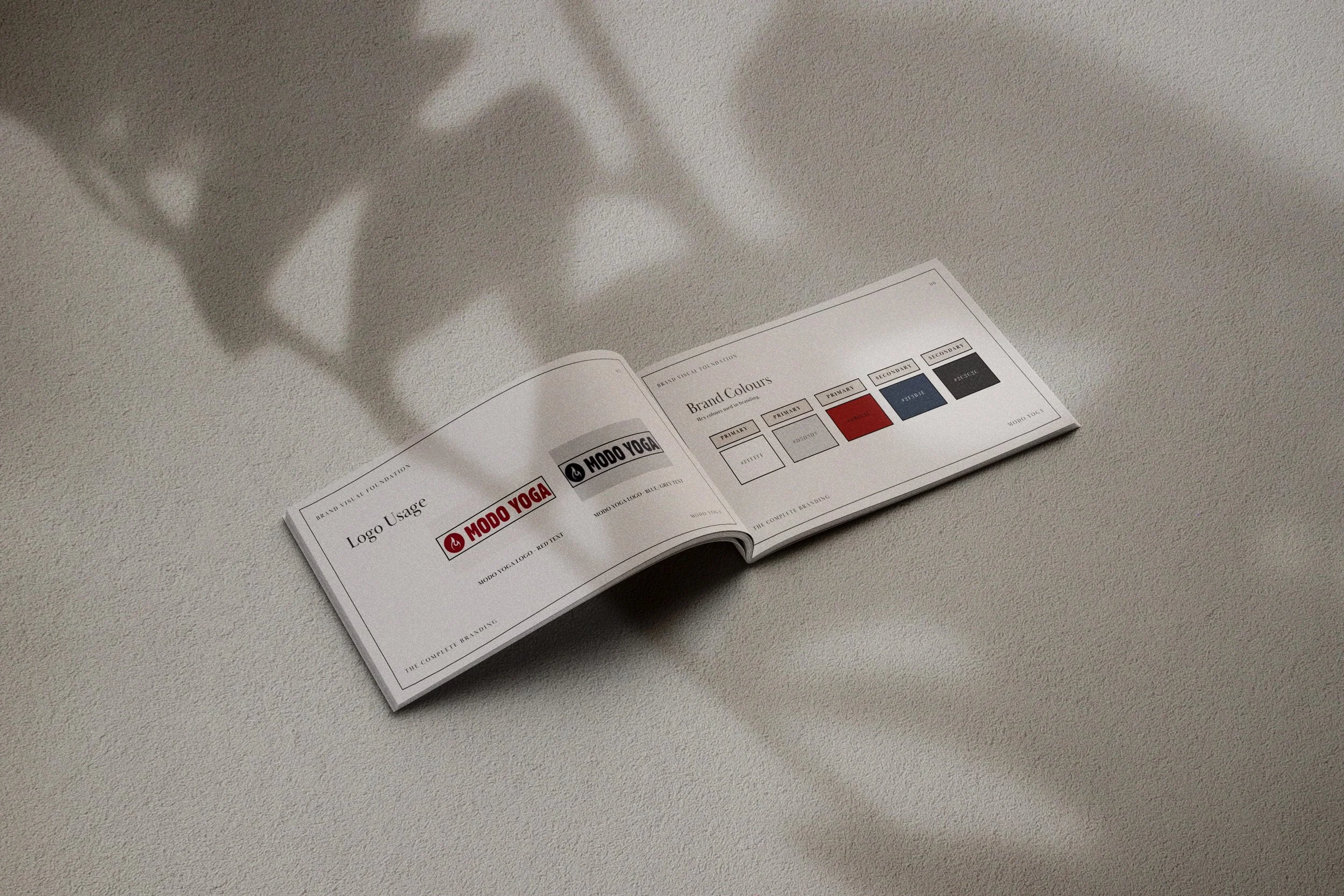 An open booklet on a light-colored textured surface displaying information about logo usage and brand colors, including red, black, blue, and grey swatches.