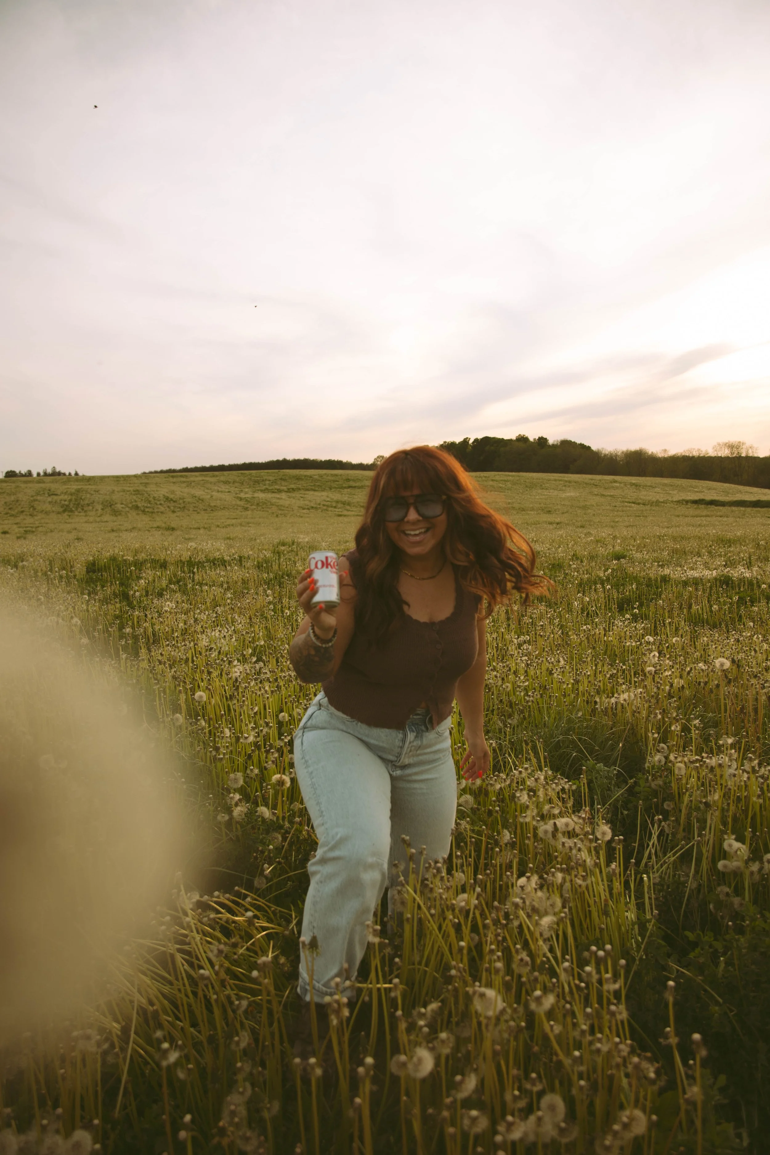 A woman with long red hair, sunglasses, and a dark top standing in a field of dandelions holding a Diet Coke can and smiling at the camera, with a grassy hill and trees in the background under a cloudy sky.