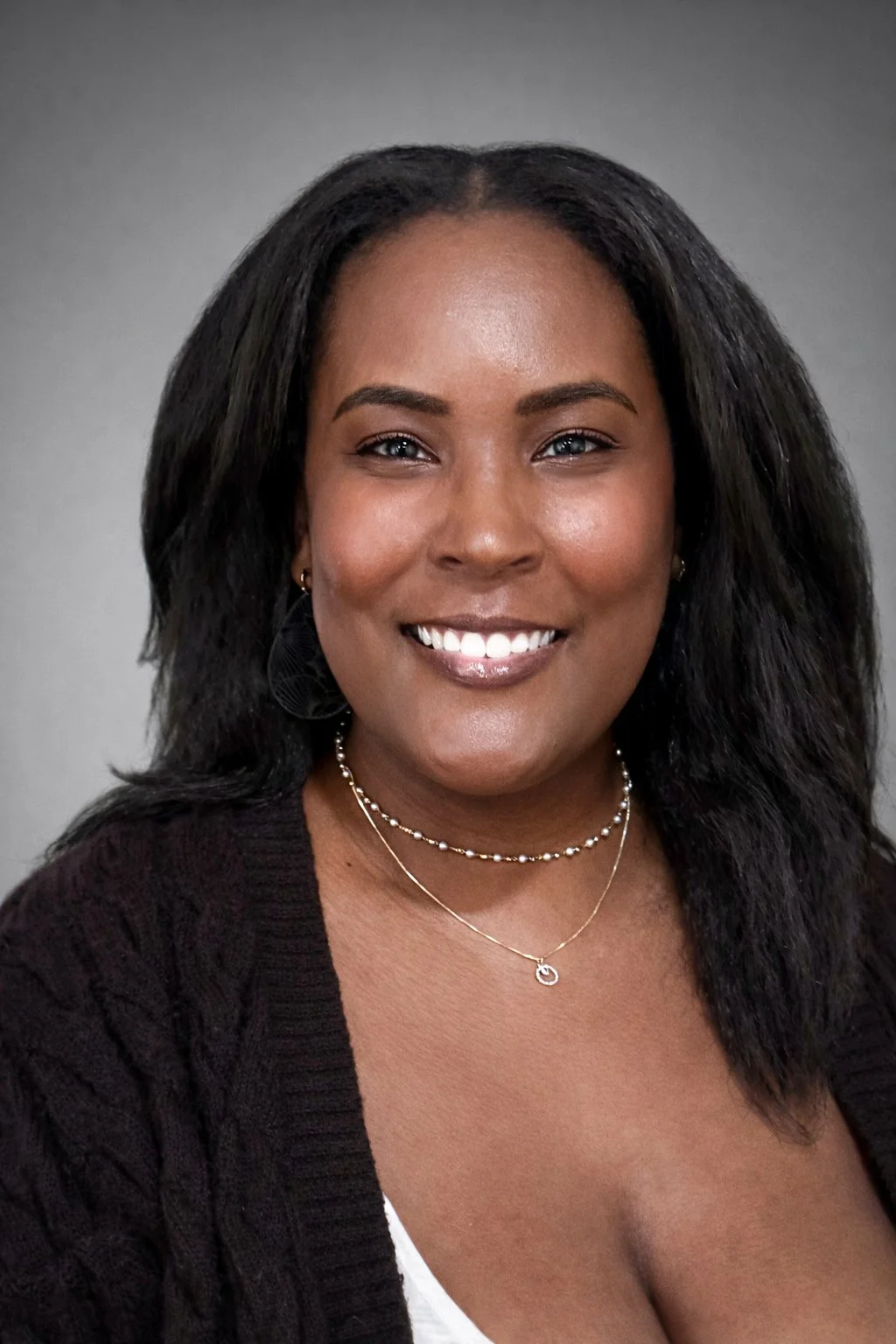 A woman with dark hair smiling at the camera, wearing a black sweater, jewelry, and a white top, in front of a neutral gray background.