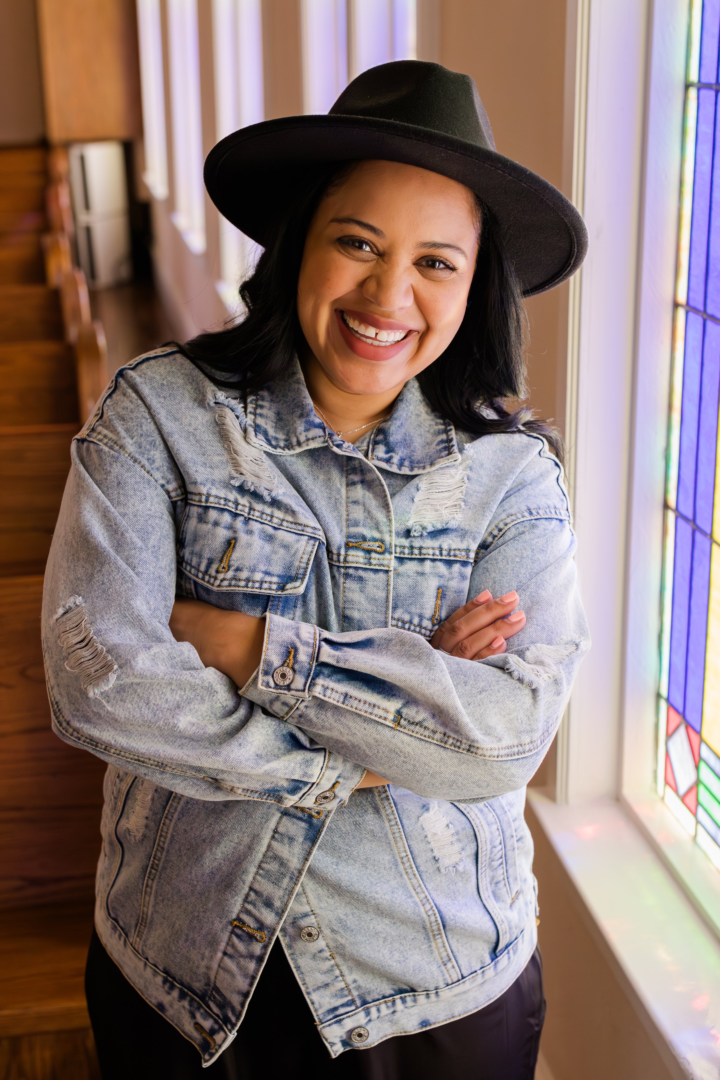 A smiling woman with black hair wearing a black wide-brimmed hat and a light blue denim jacket, standing by a stained glass window in a warmly lit room.