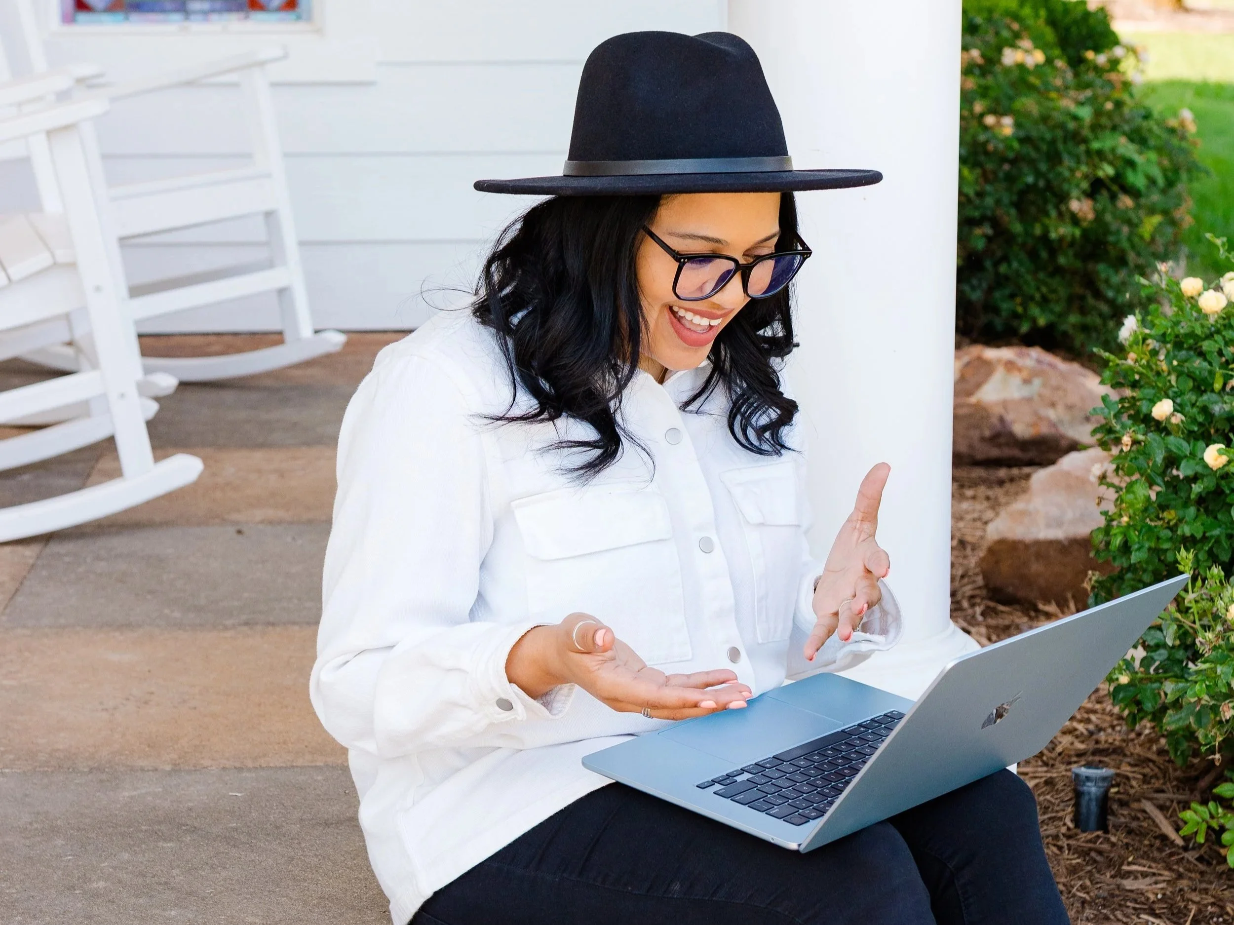A woman with black hair, glasses, and a black hat sitting outside on a bench with a white jacket, smiling and gesturing while looking at her laptop.