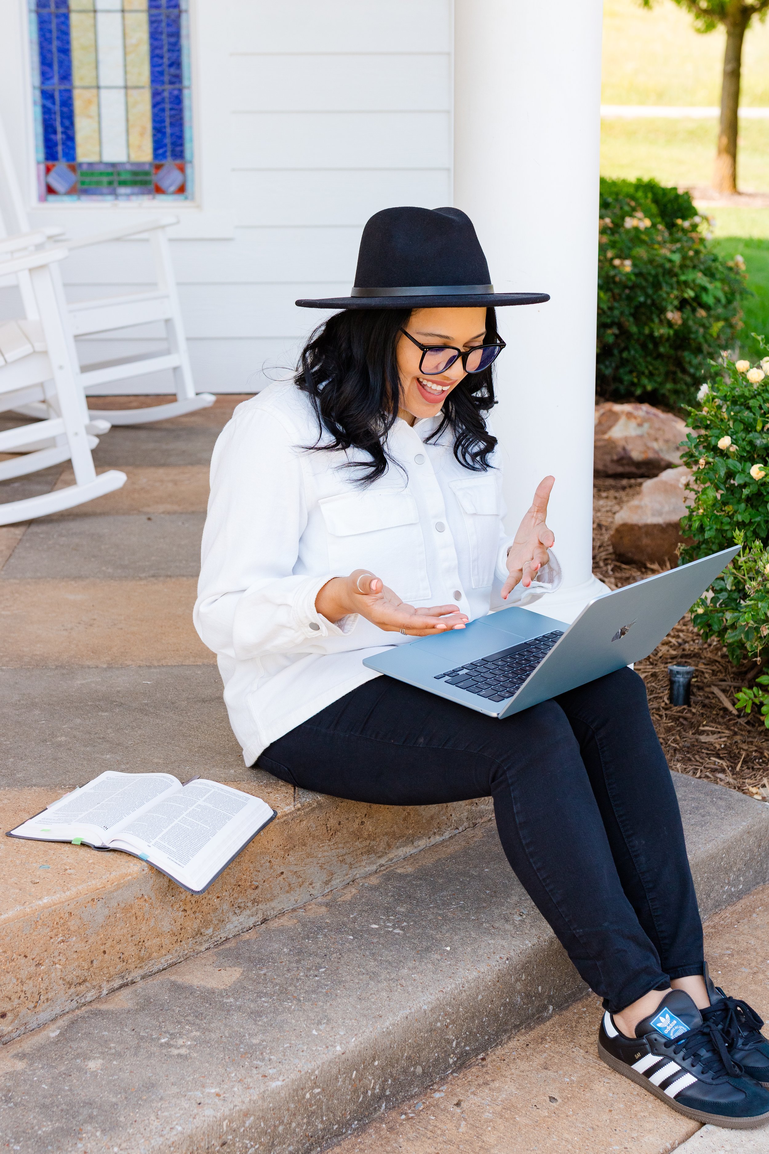 A woman with black hair, glasses, and a black hat sits outside on a concrete step, smiling and using a silver laptop. There are open books on the ground nearby, and she is dressed in a white shirt and black pants. The background includes a white house with colorful stained glass window, a white rocking chair, and green bushes and trees.