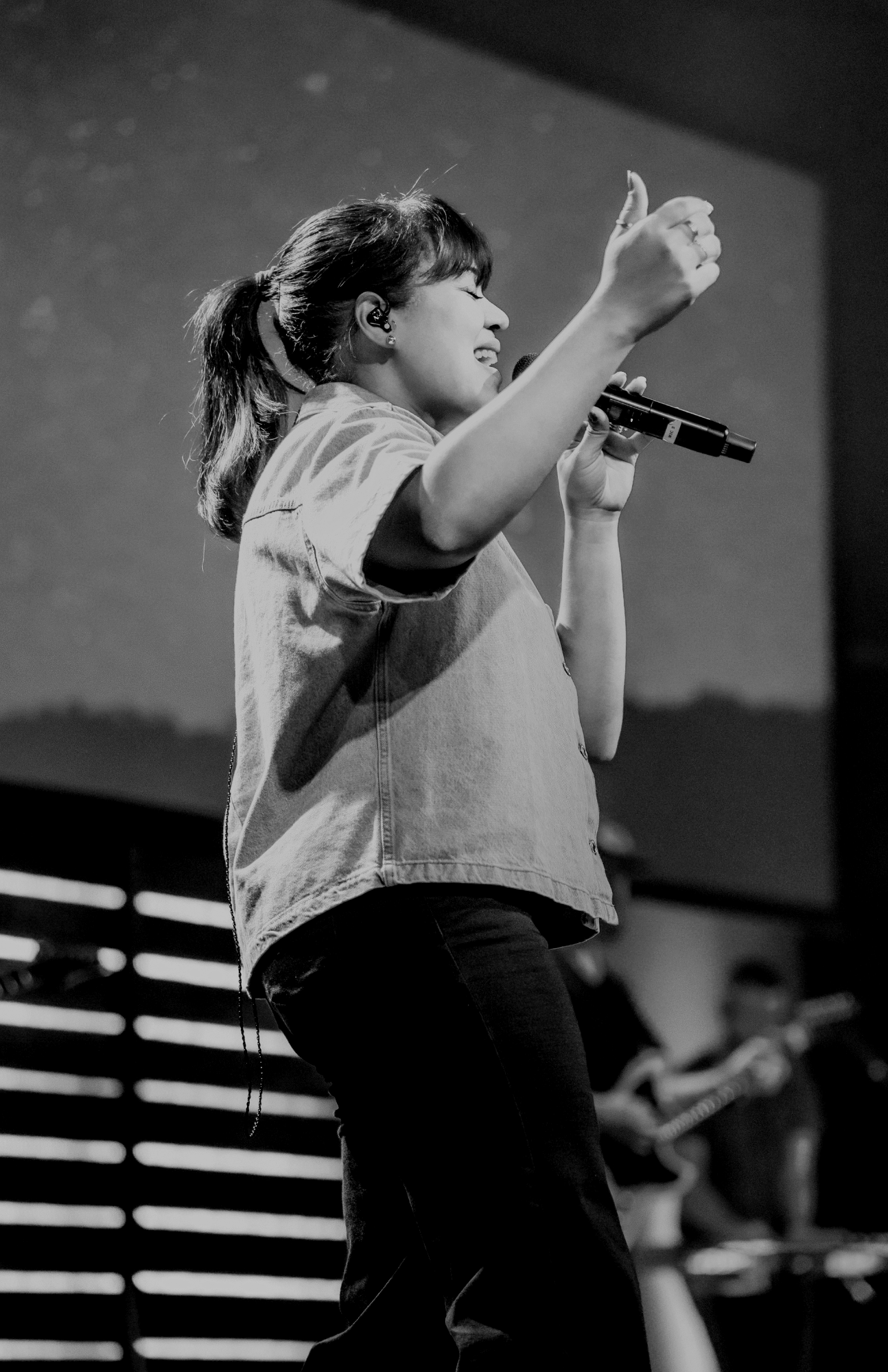 A woman singing into a microphone, with her eyes closed and one arm raised, on a stage with a background of horizontal lines.