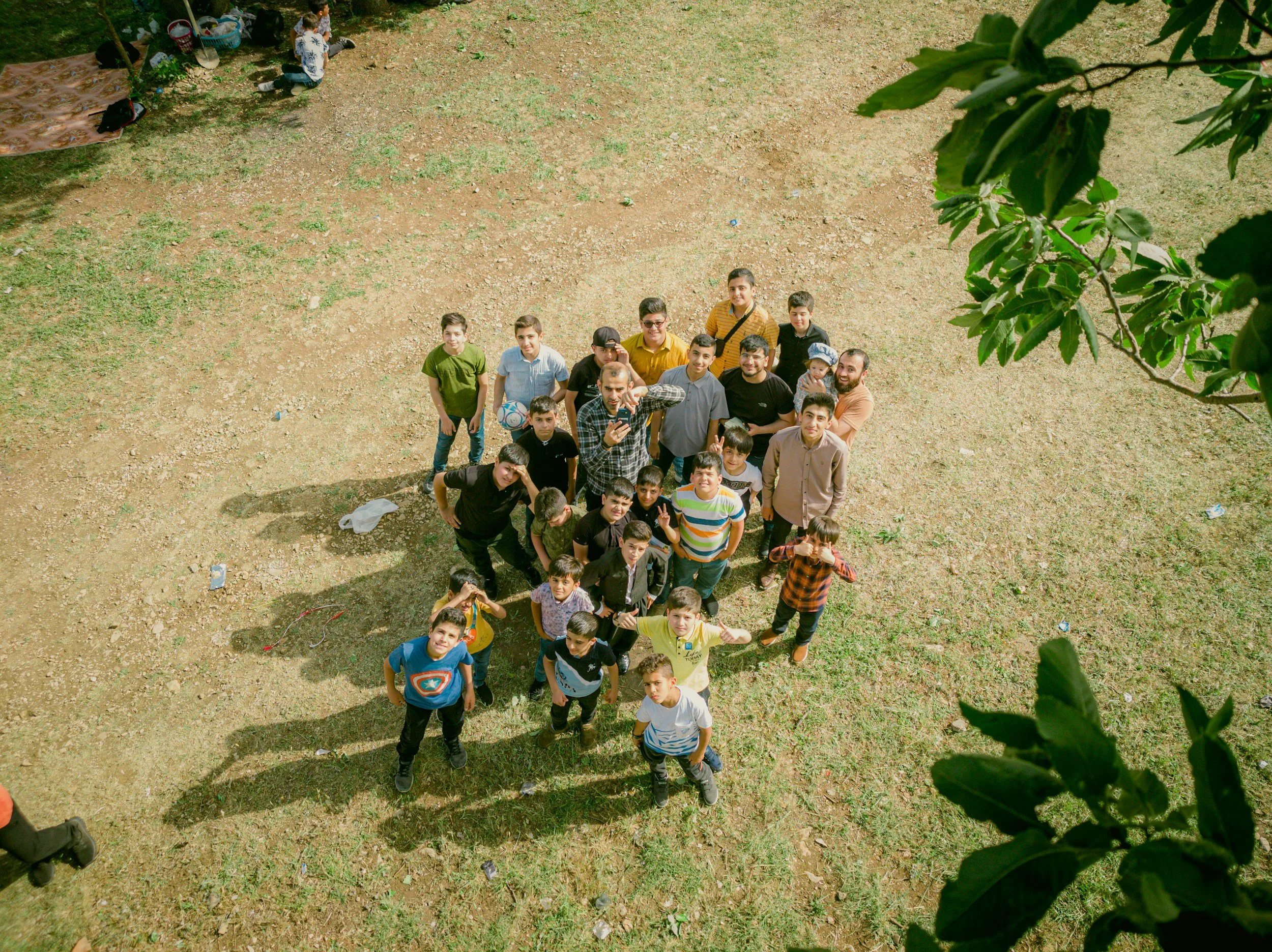 A group of children and young adults gathered outdoors on a dirt and grass area, looking up at the camera taking a selfie from above, with trees and a blanket in the background.
