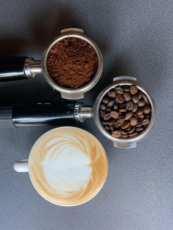 A cup of latte with foam art, two containers of ground coffee and coffee beans, all on a dark gray surface.