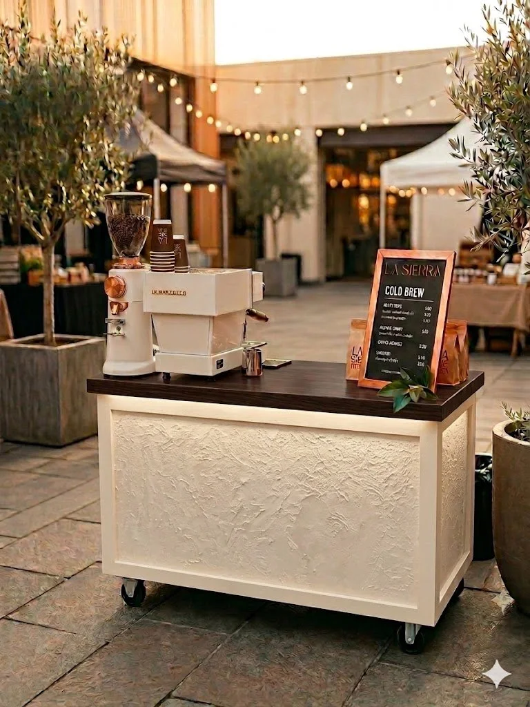 A portable coffee station with a white textured base and a dark wooden top, set up outdoors at a café or event. There is a coffee grinder and a hot beverage machine on the counter, along with a blackboard menu listing cold brew options and prices. Potted trees and string lights are visible in the background, creating a cozy atmosphere.
