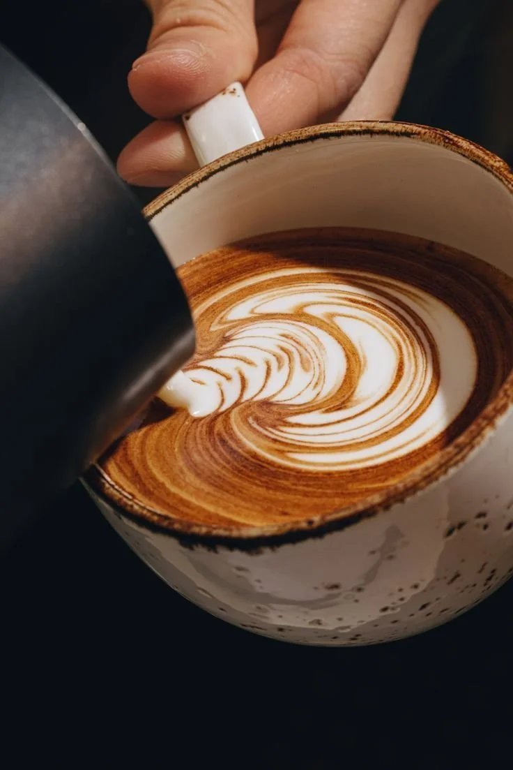 A barista is pouring steamed milk into a cup of espresso, creating latte art on the surface.
