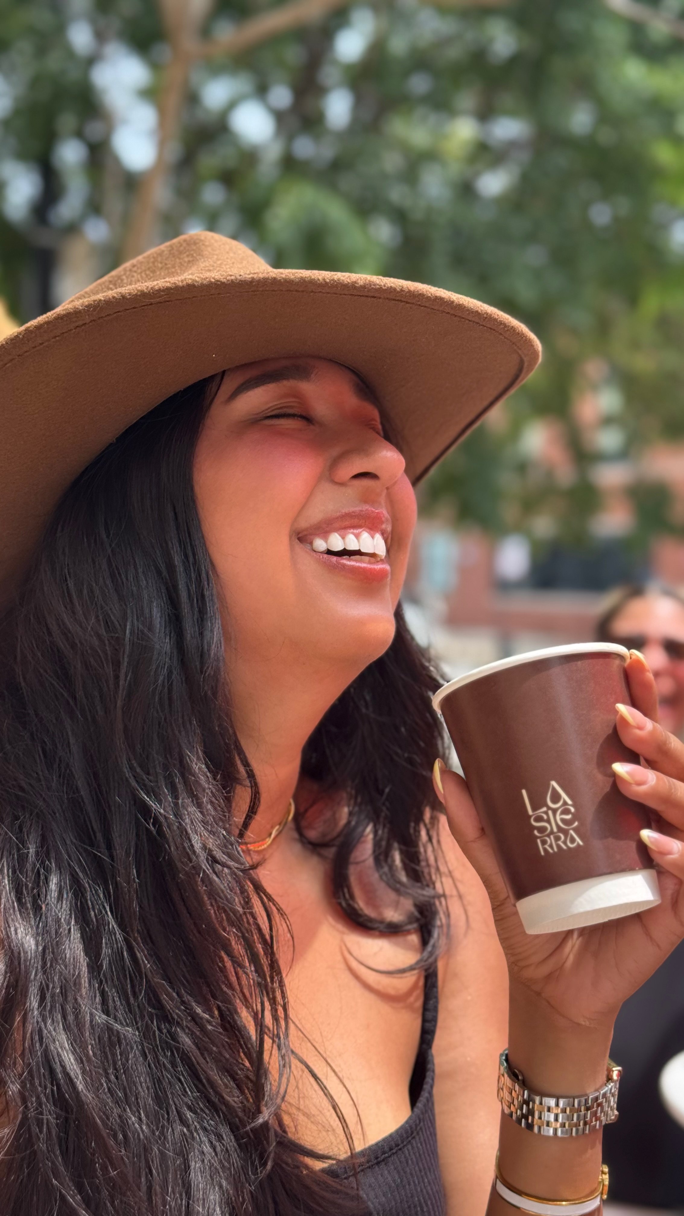 Woman wearing a wide-brimmed tan hat, holding a coffee cup, smiling and laughing outdoors with blurred green trees in the background.