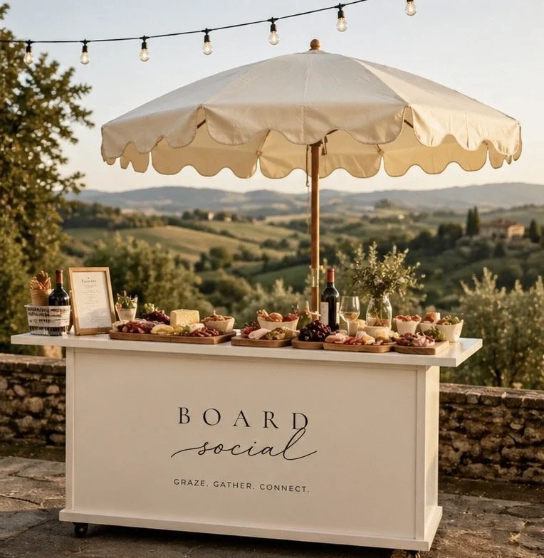Outdoor table setup with an umbrella, wine bottles, wine glasses, cheese, meats, grapes, and snacks, set against a scenic countryside view with string lights overhead.
