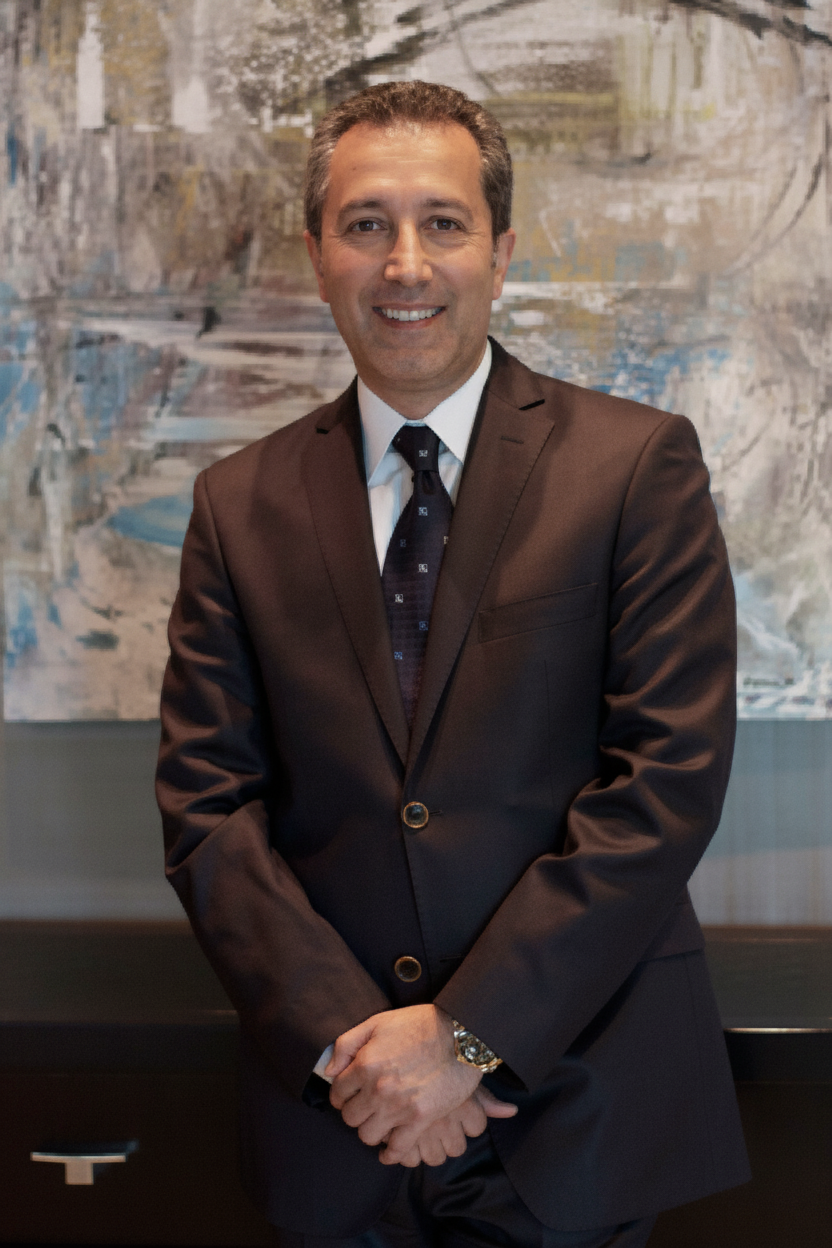 A man in a brown suit with a white shirt and a dark patterned tie, standing with his hands clasped in front of him, smiling at the camera.