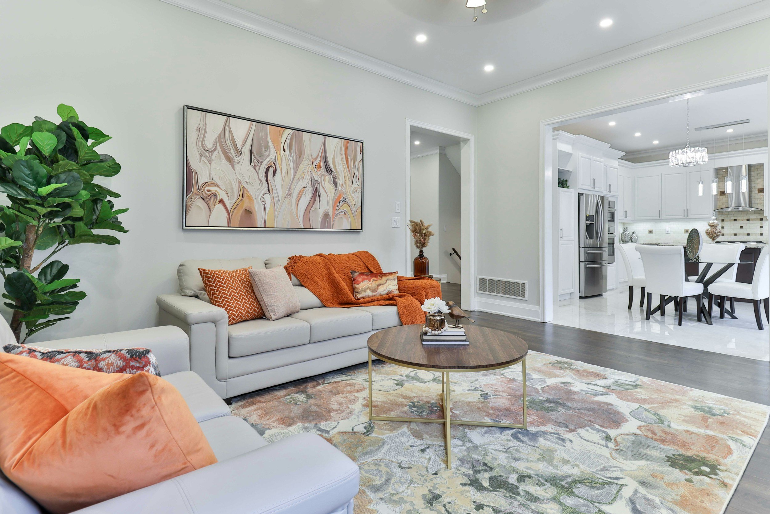 Living room with white sofa, orange throw blanket, decorative pillows, a round wooden coffee table, large abstract art on the wall, green houseplant, and an adjoining kitchen with white cabinets, stainless steel appliances, and a chandelier.