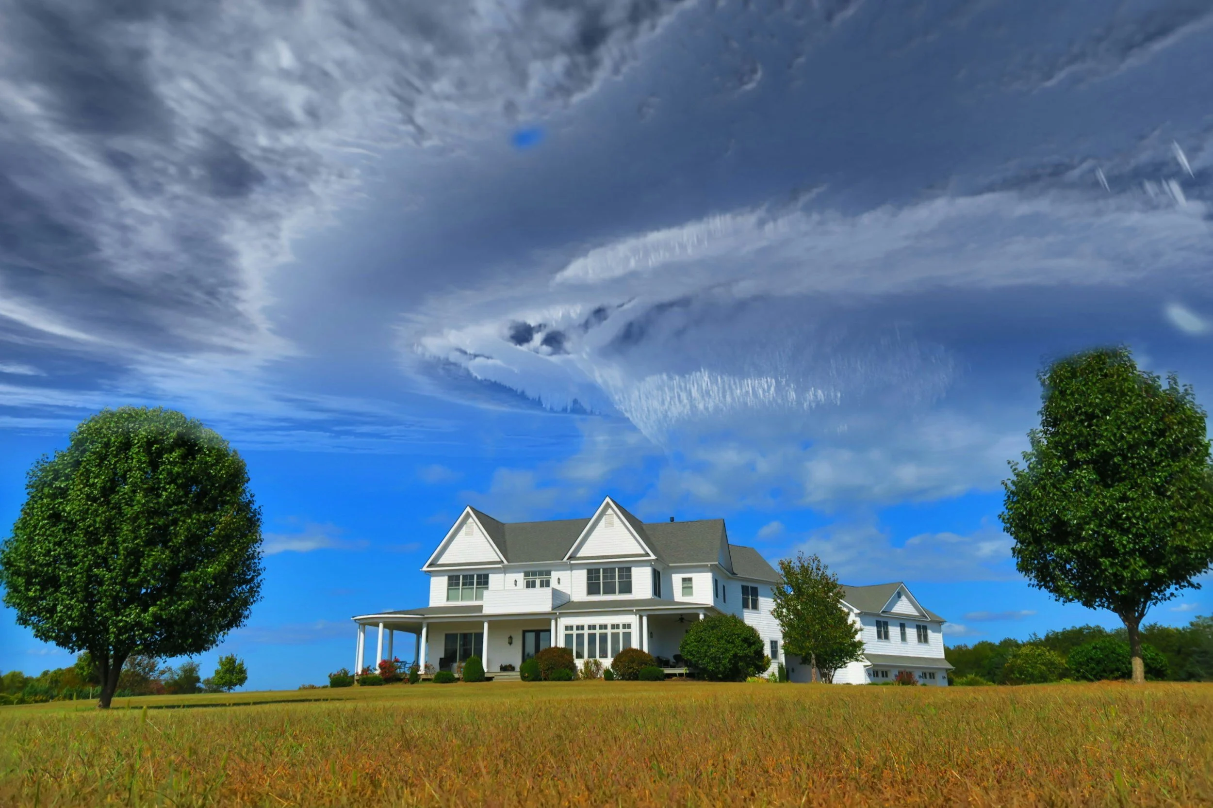 A white multi-story house with a wraparound porch, surrounded by green grass and trees, under a blue sky with large, dramatic clouds.