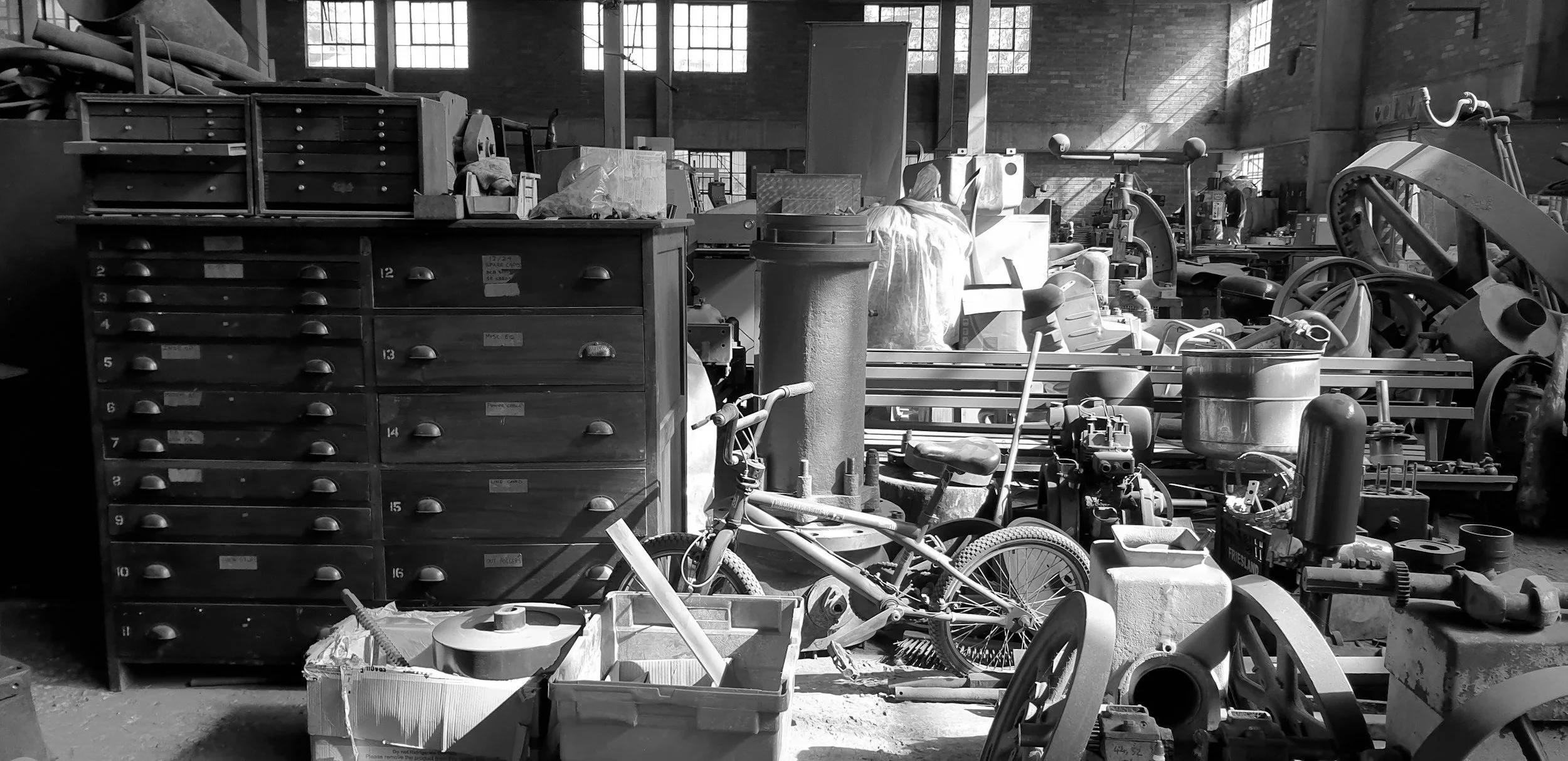 A cluttered workshop with tools, boxes, a bicycle, and heavy machinery in a brick building with large windows.