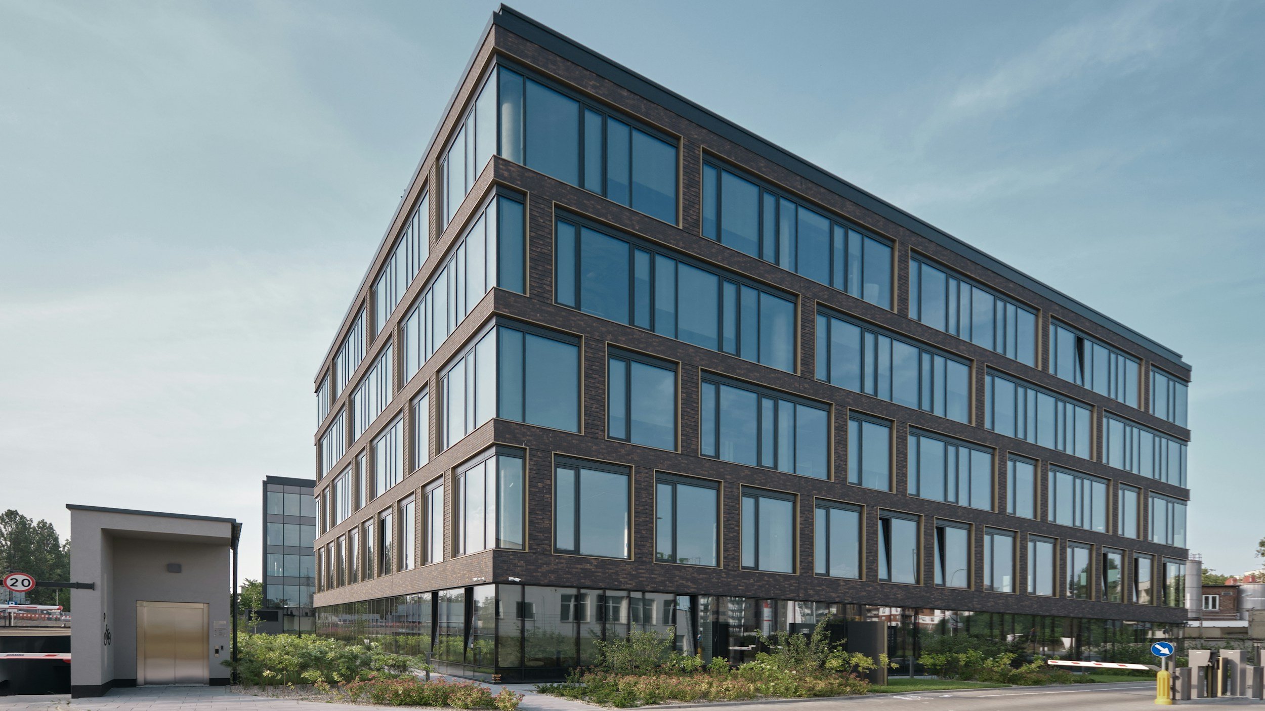 Modern multi-story building with large glass windows and dark brick exterior, situated in an urban area under a partly cloudy sky.