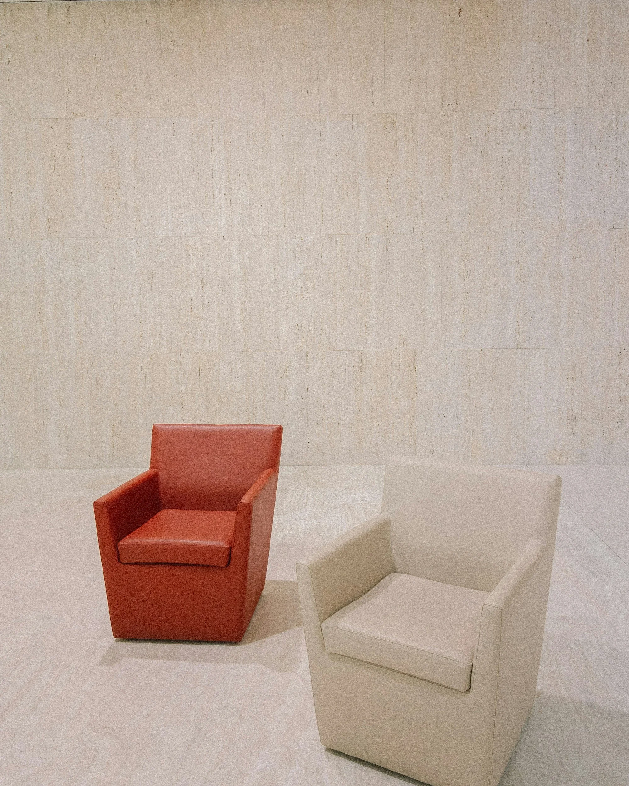 Two modern armchairs, one red and one beige, placed on a light-colored floor against a plain wall.