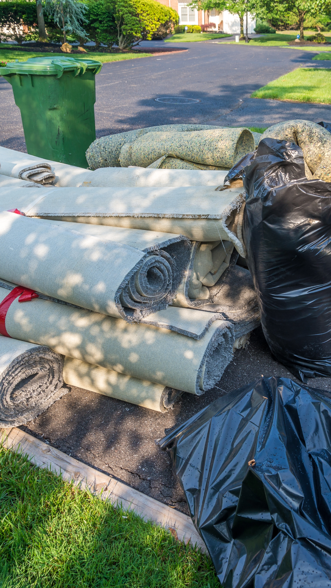 Trimmed and rolled carpets, a black garbage bag, and a green trash bin placed on asphalt near a grassy area with trees and houses in the background.