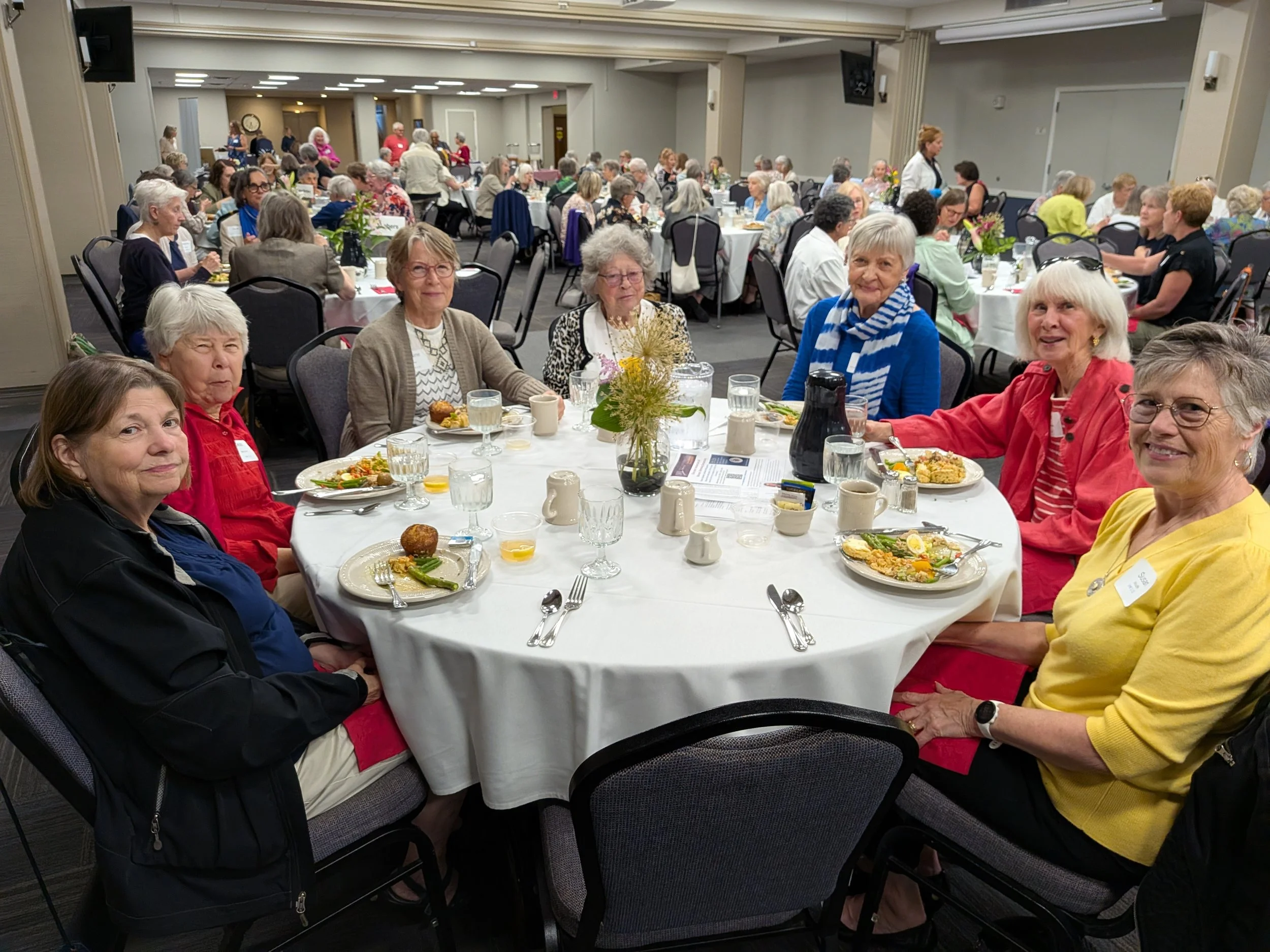A large group of elderly women sitting around several round tables at a banquet or gathering, enjoying a meal together in a conference or community center.