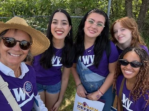 Group of five women smiling outdoors, wearing purple shirts with white text, one woman with a large hat and sunglasses, others with glasses and colorful hair, holding a sign that says 'Women Defense' amid trees in the background.