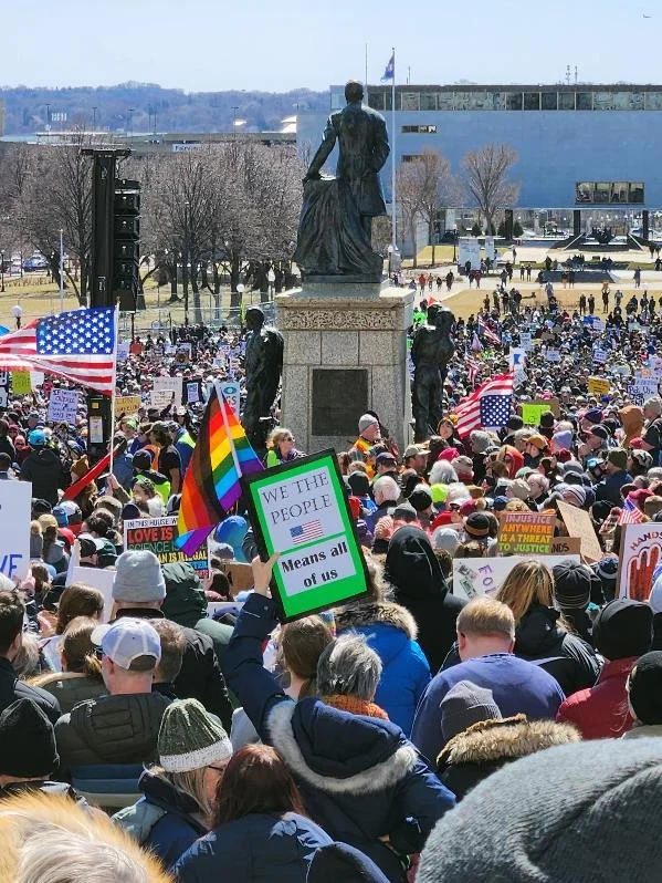Crowd gathered at a protest or rally, with many holding signs and American flags, in front of a statue and a modern building in the background.
