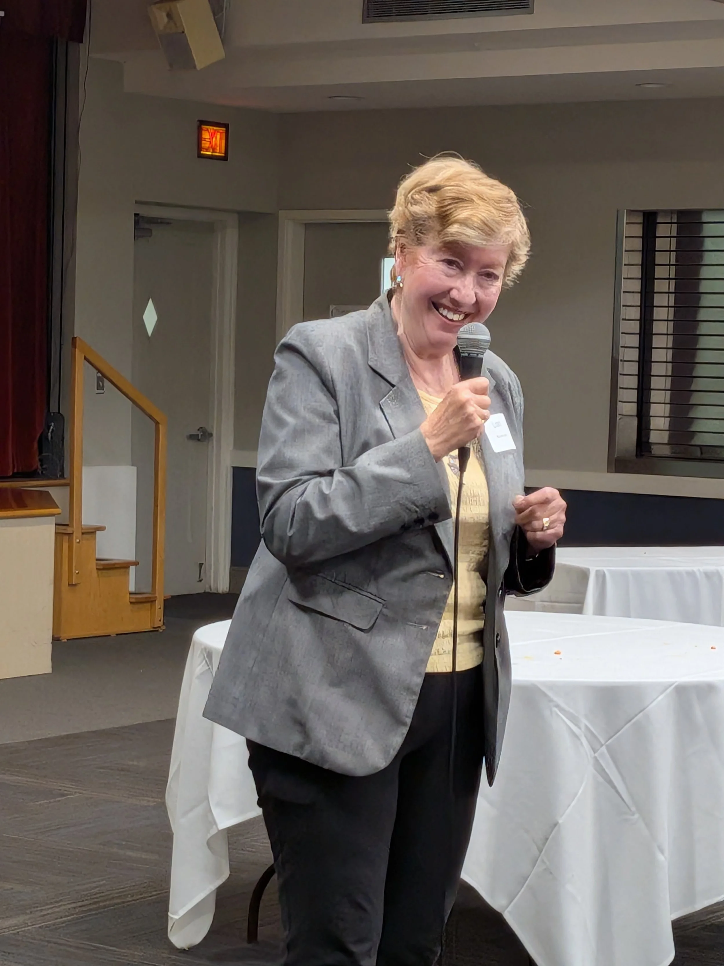 An elderly woman with short, wavy blonde hair, wearing a grey blazer and a yellow top, holding a microphone and smiling while speaking at an indoor event. Behind her are white tables with white tablecloths, and a background with doors, wall decor, an