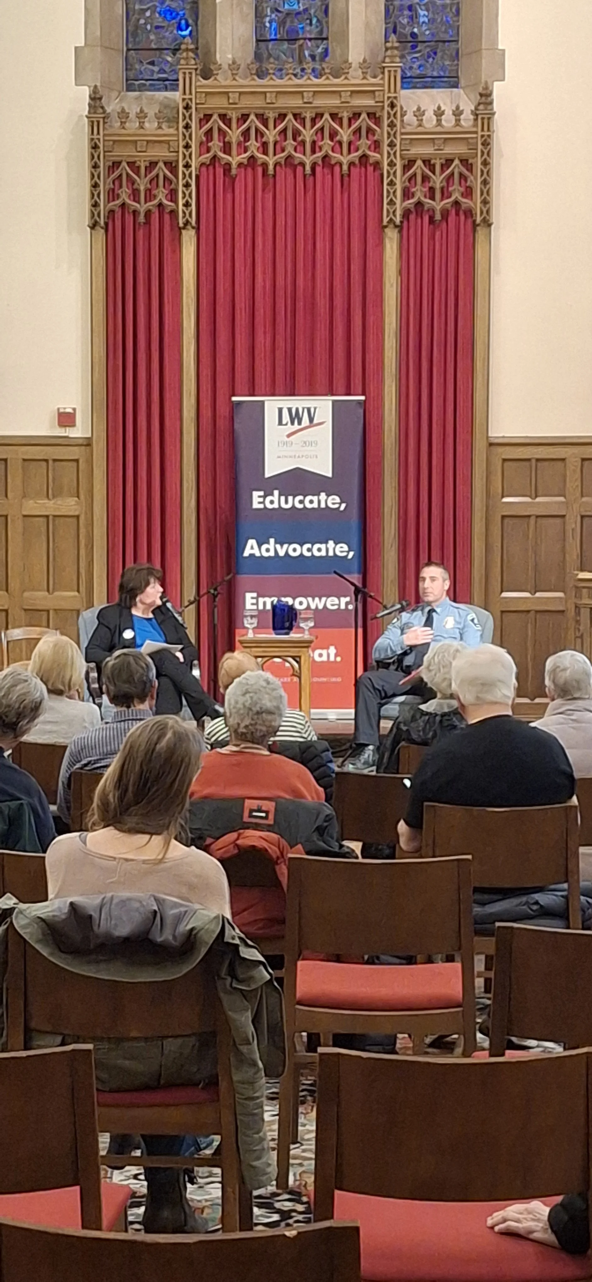 A panel discussion taking place in a wood-paneled room with red curtains. Two speakers, a woman and a man, sit on stage with microphones. Audience members are seated, listening. A vertical banner reads 'LWV 1919-2019 Educate, Advocate, Empower'.