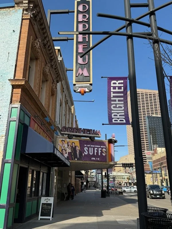 Street view of Orpheum Theater with vertical sign and purple banner reading 'BRIGHTER'. The theater has a marquee advertising the show 'SUFFS' from April 7-12, with a billboard featuring a cast of actors. The scene is in daylight with a clear blue sk