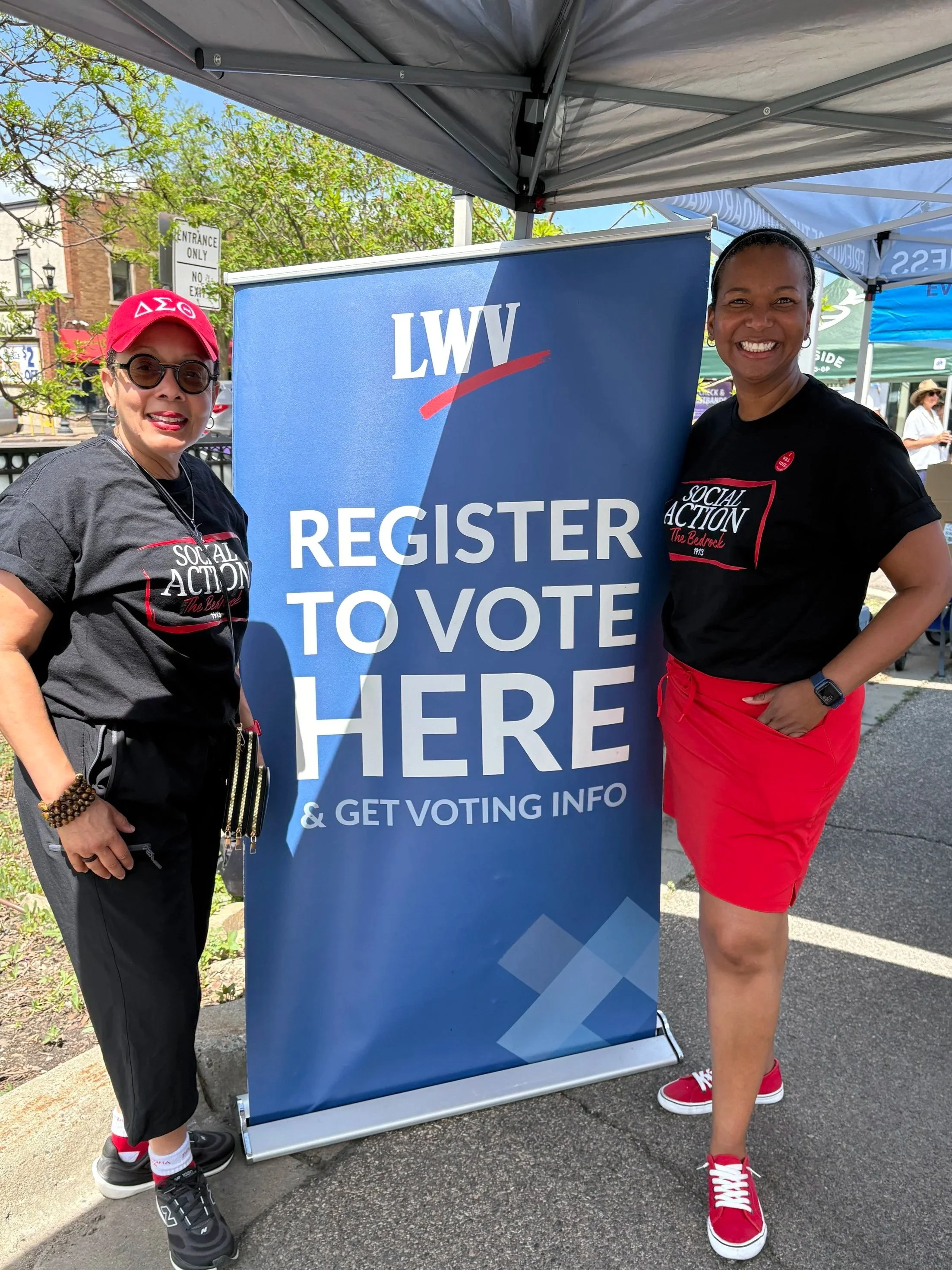 Two women standing next to a blue sign that reads 'Register to Vote Here' at a voting registration event. They are smiling and wearing black t-shirts with white and red text, and red and black sneakers. One woman is wearing sunglasses and a red baseb
