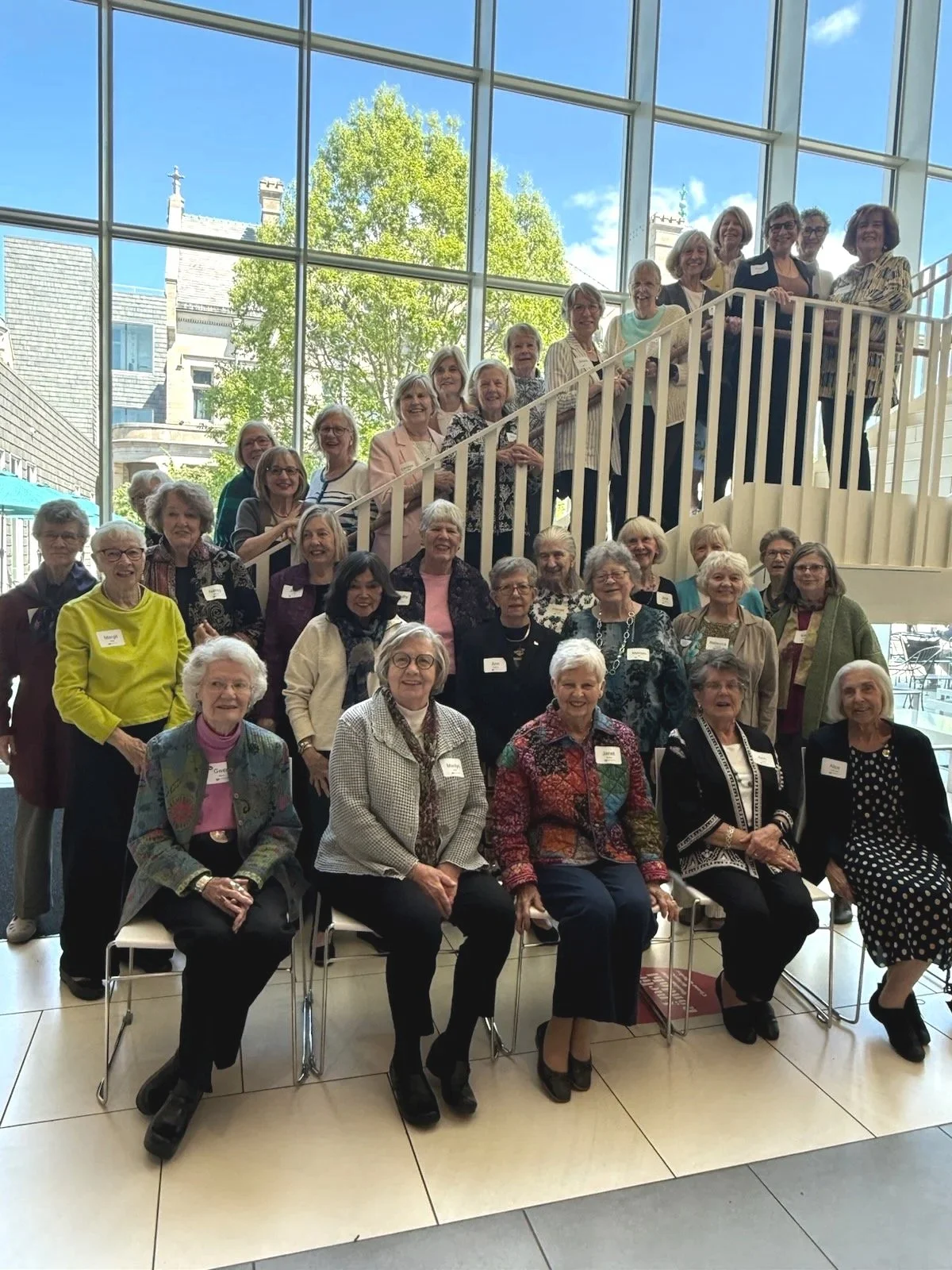 Group of elderly women gathered in a brightly lit indoor space with large windows and a modern staircase, some seated and some standing, smiling for a group photo.