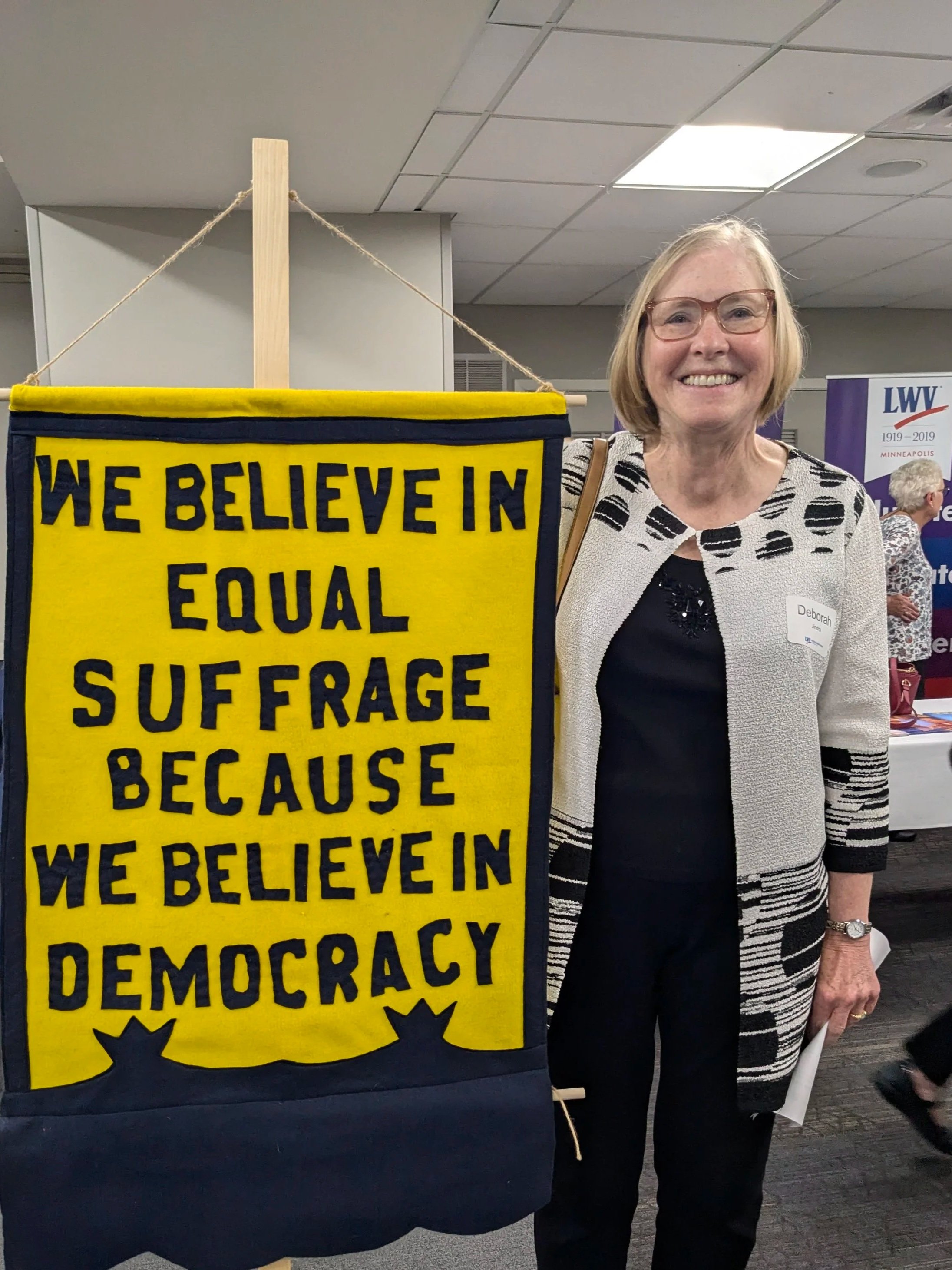 A smiling woman wearing glasses and a patterned jacket standing next to a yellow and navy blue banner that reads, "We believe in equal voice because we believe in democracy." In the background, there are other people at an indoor event with banners a