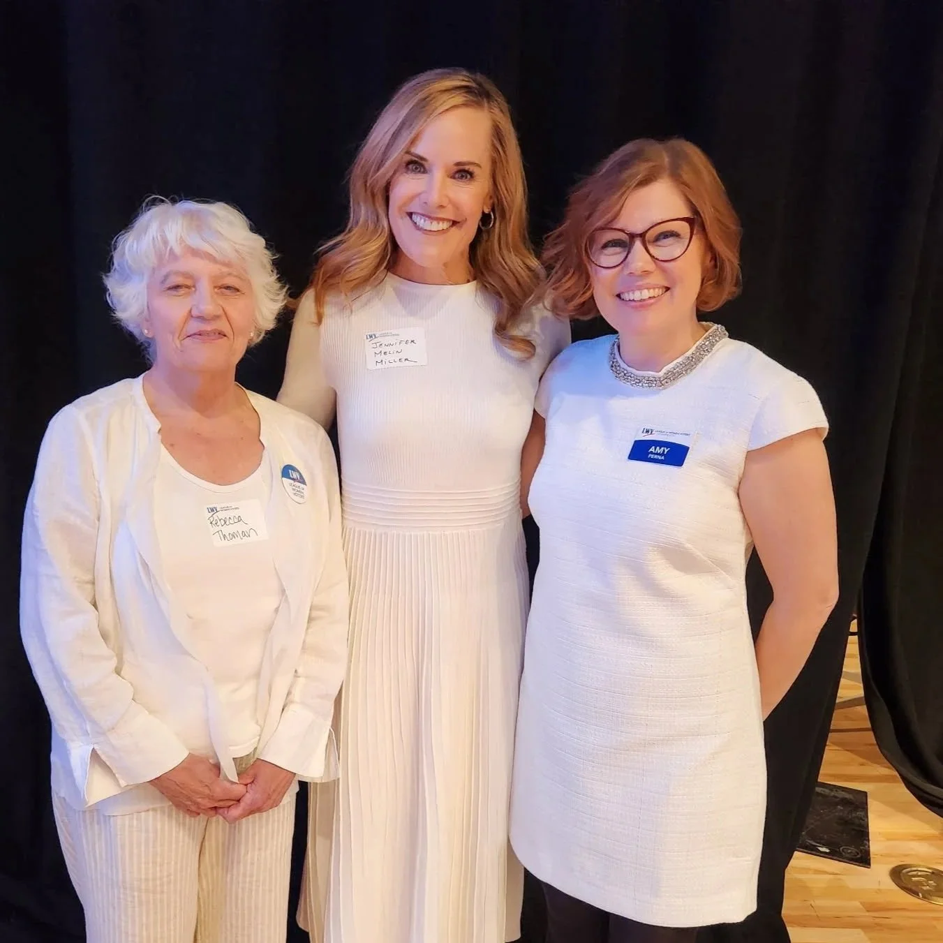 Three women standing together at an event, all wearing white outfits and name tags, with a dark backdrop behind them.
